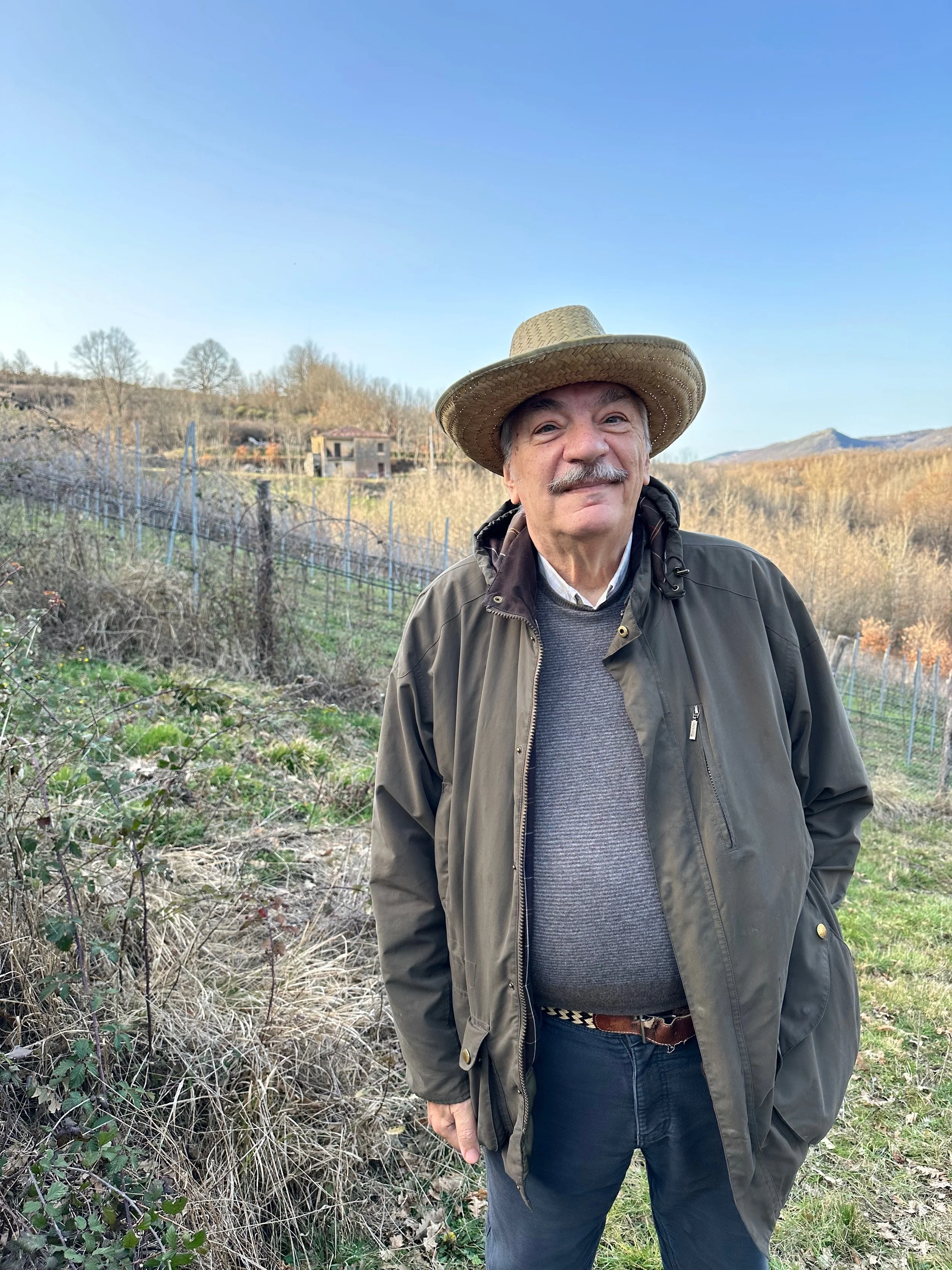 Uomo anziano con cappello di paglia e giacca scura che sorride in un paesaggio rurale con campi, alberi e montagne sullo sfondo.