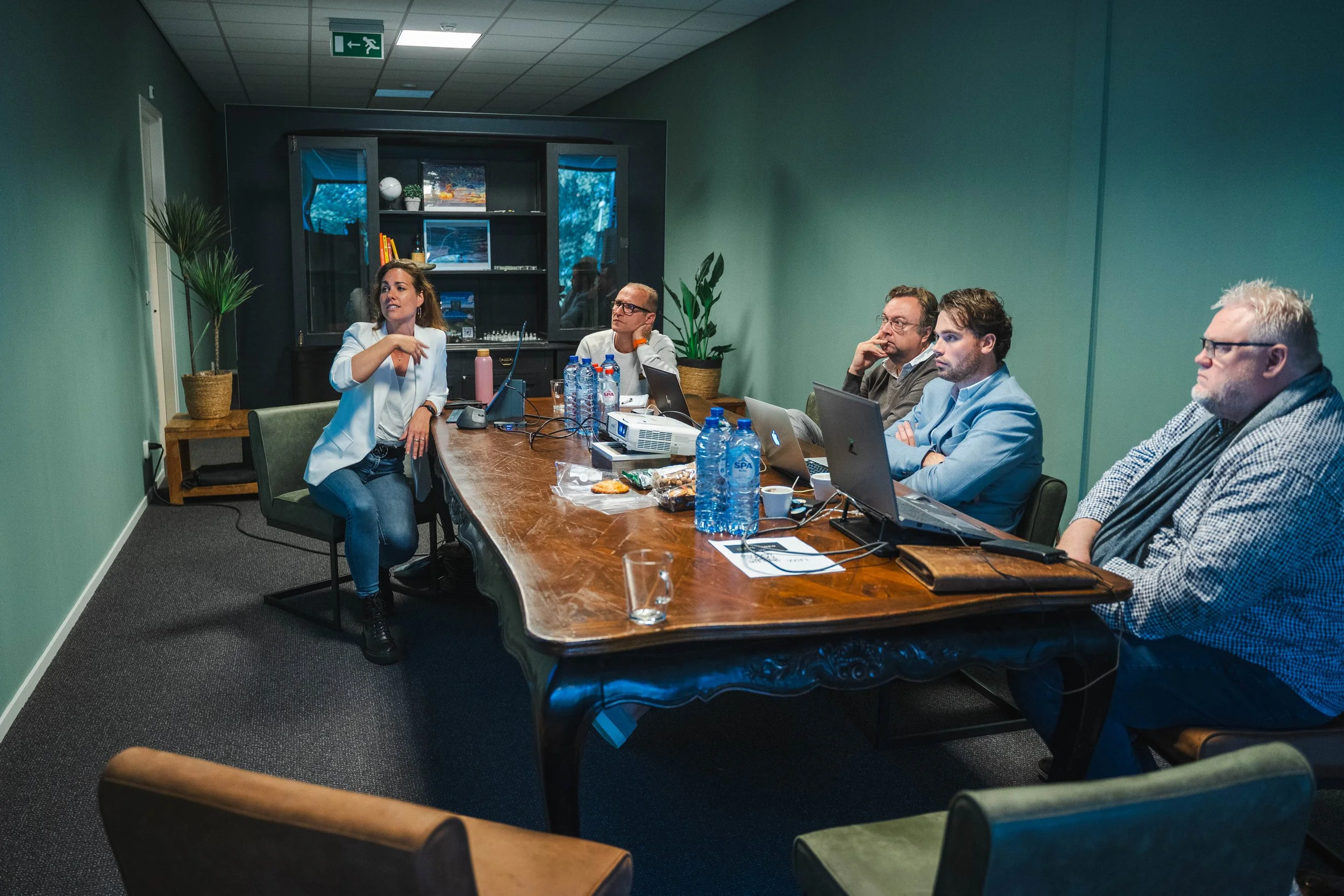 Five people sitting around a table with laptops and water bottles, having a discussion in a meeting room.
