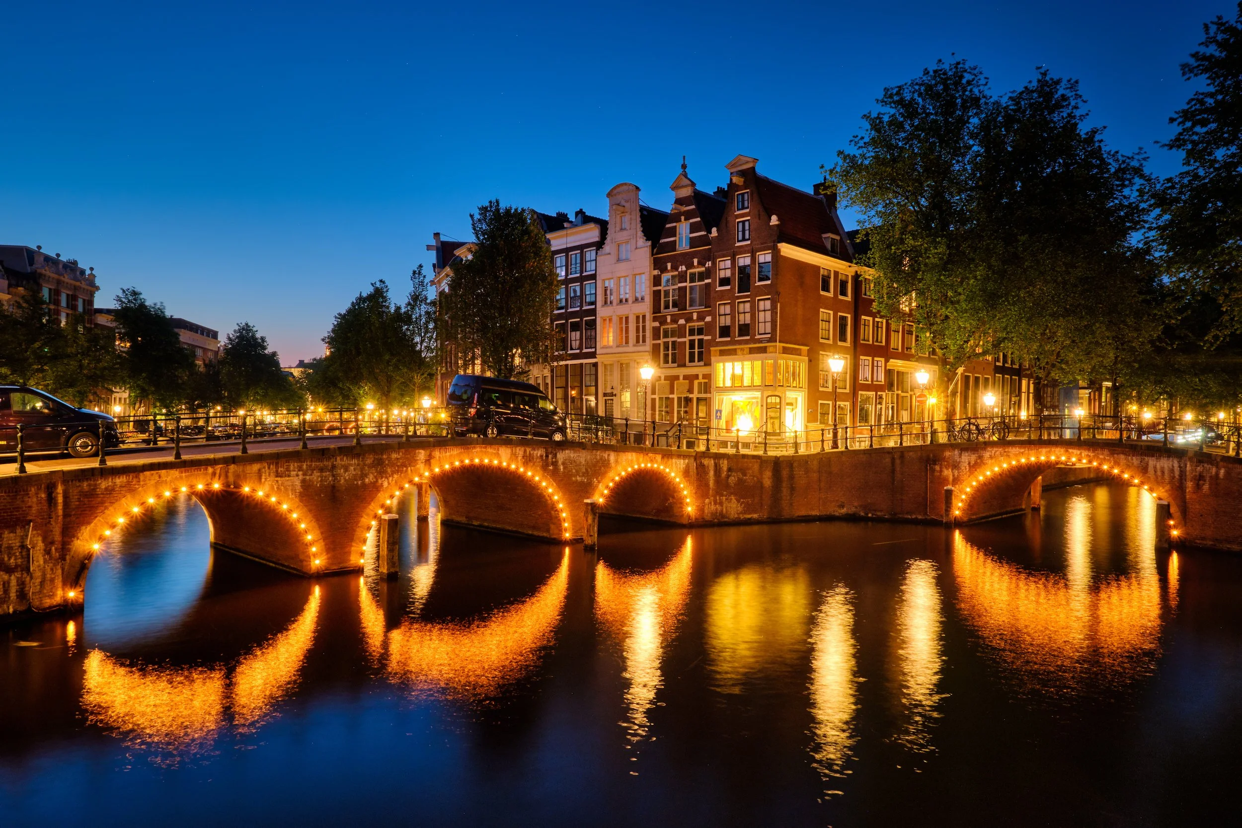 Night view of an illuminated canal bridge in Amsterdam with historic buildings and trees reflecting in the water.