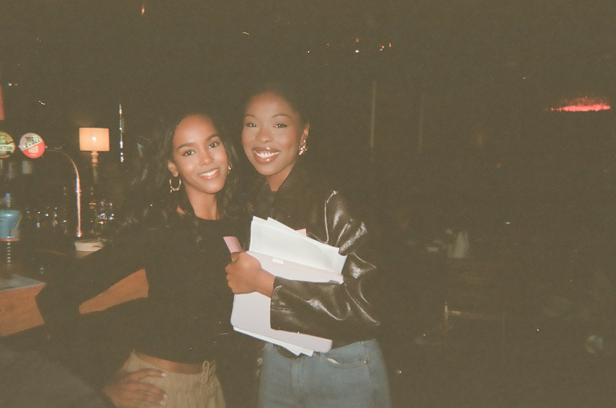 Two women smiling and taking a selfie inside a dimly lit venue, one holding some papers or booklets, with a few tables and wall decorations visible in the background.