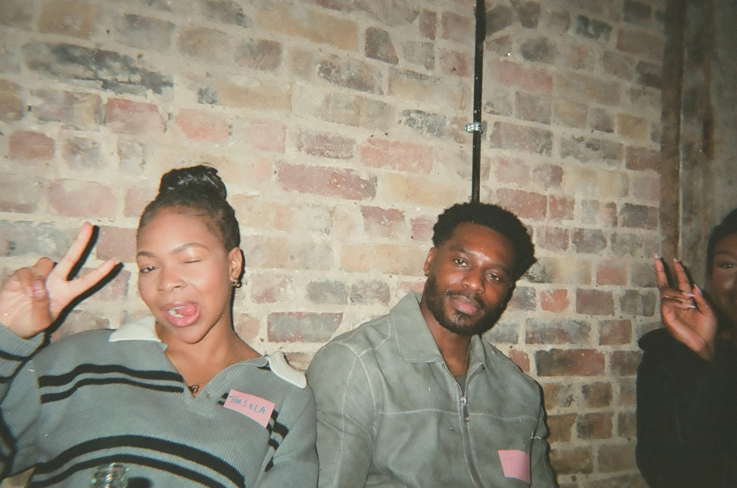 Three young adults posing in front of a brick wall, making peace signs with their hands. One woman is winking and sticking her tongue out, while the other two appear relaxed. They are casually dressed, and one has a name tag that says Sola.