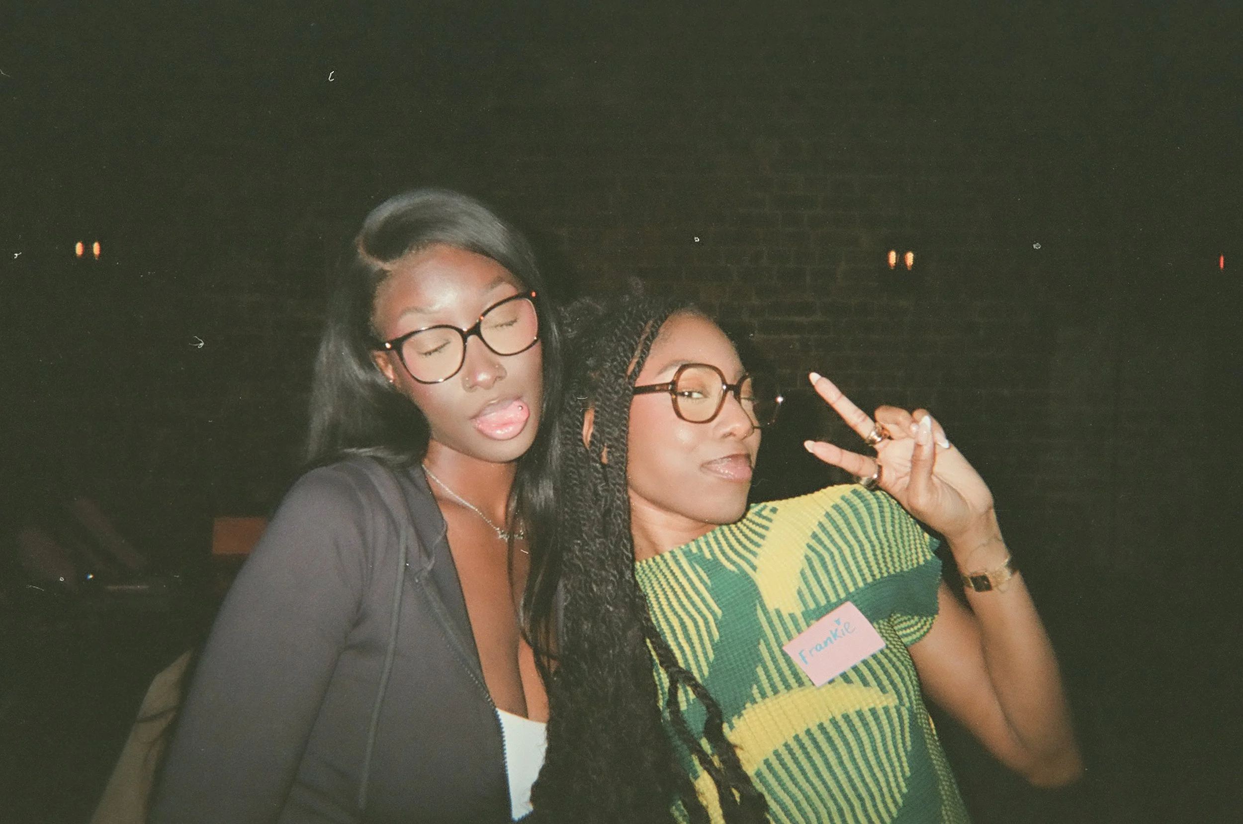 Two women posing together at an indoor event, one with long dark hair, glasses, sticking tongue out, and the other with braided hair, glasses, making a peace sign, wearing a colorful striped shirt with a name tag that reads "Frankie".