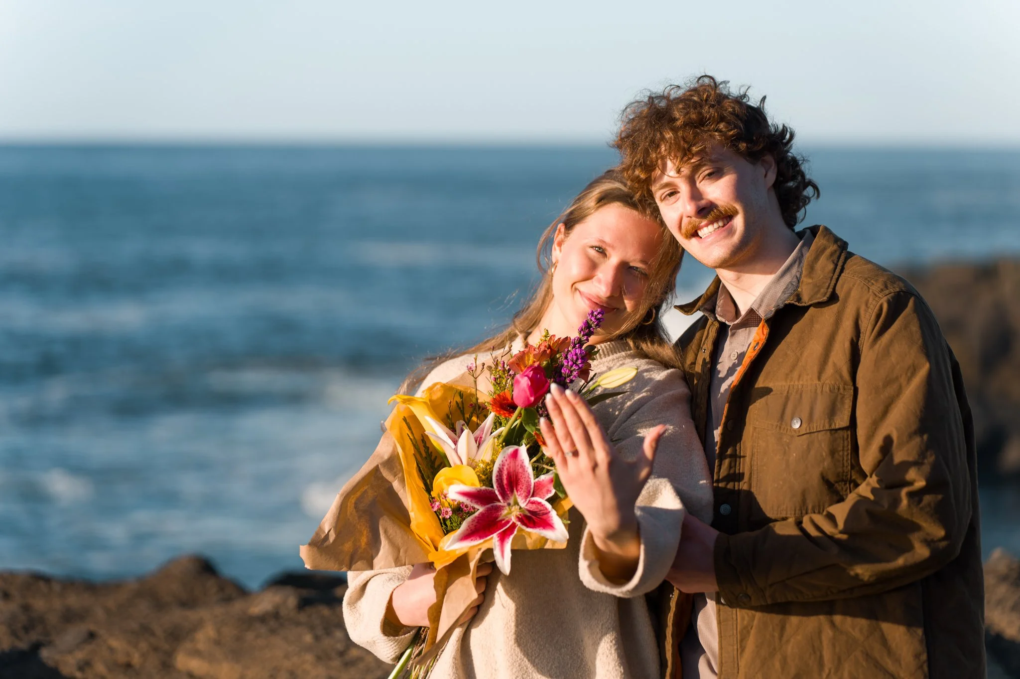 A couple posing for the camera with their bouquet of flowers and a diamond ring
