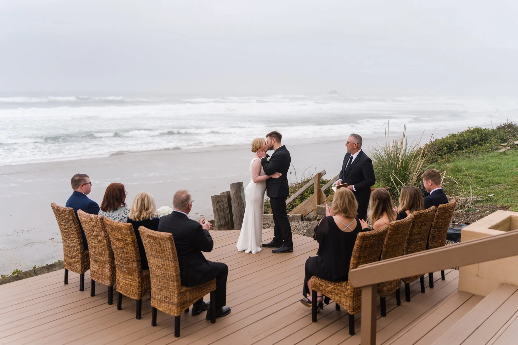 Bride and groom share their first kiss during a micro wedding on a beach-view Airbnb deck in Lincoln City, Oregon, overlooking Roads End Beach with God’s Thumb in the background as family watches.
