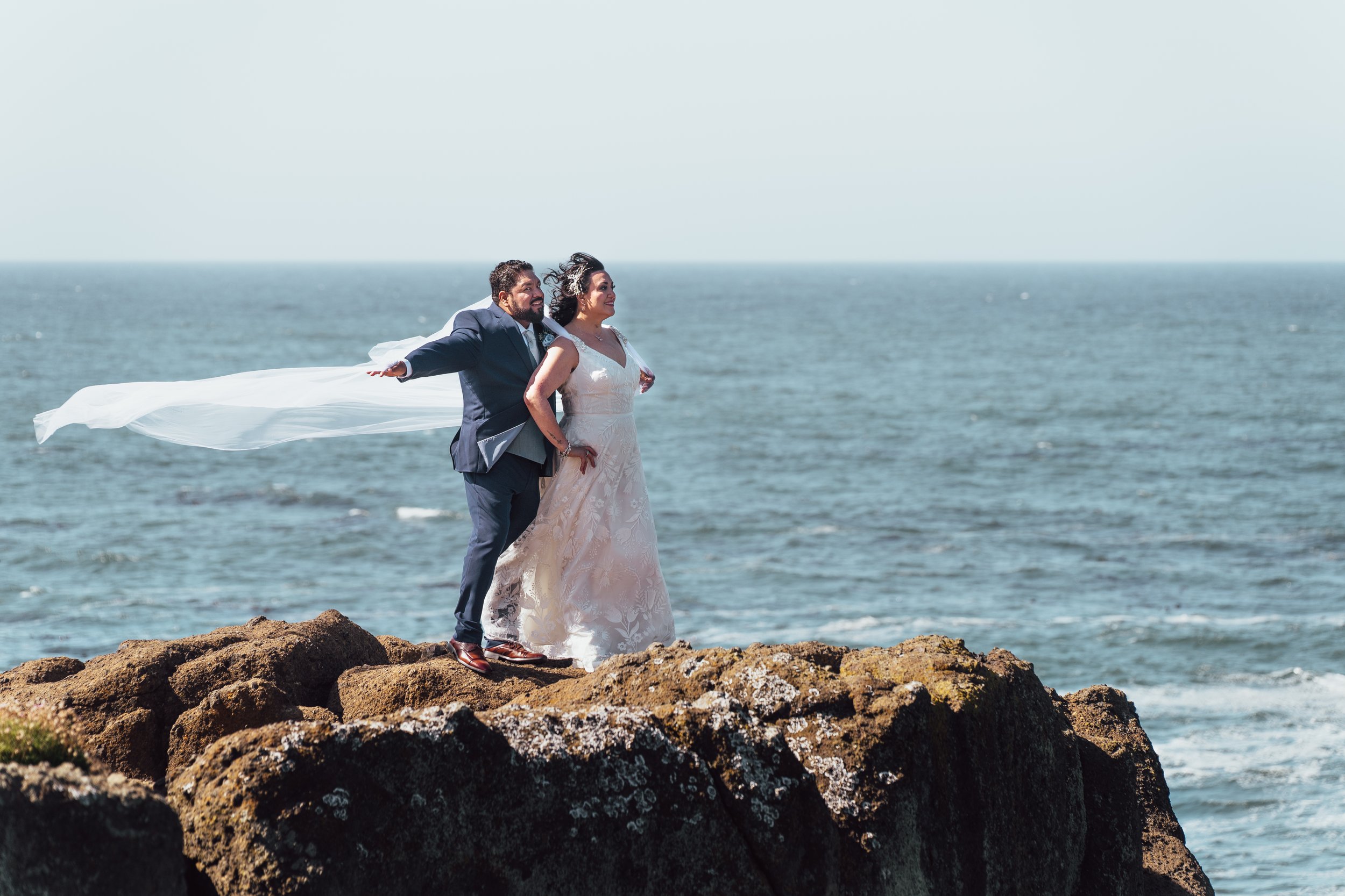 A bride and groom in wedding attire standing on rocks by the ocean, with the groom's jacket open and a white cloth blowing in the wind behind them.