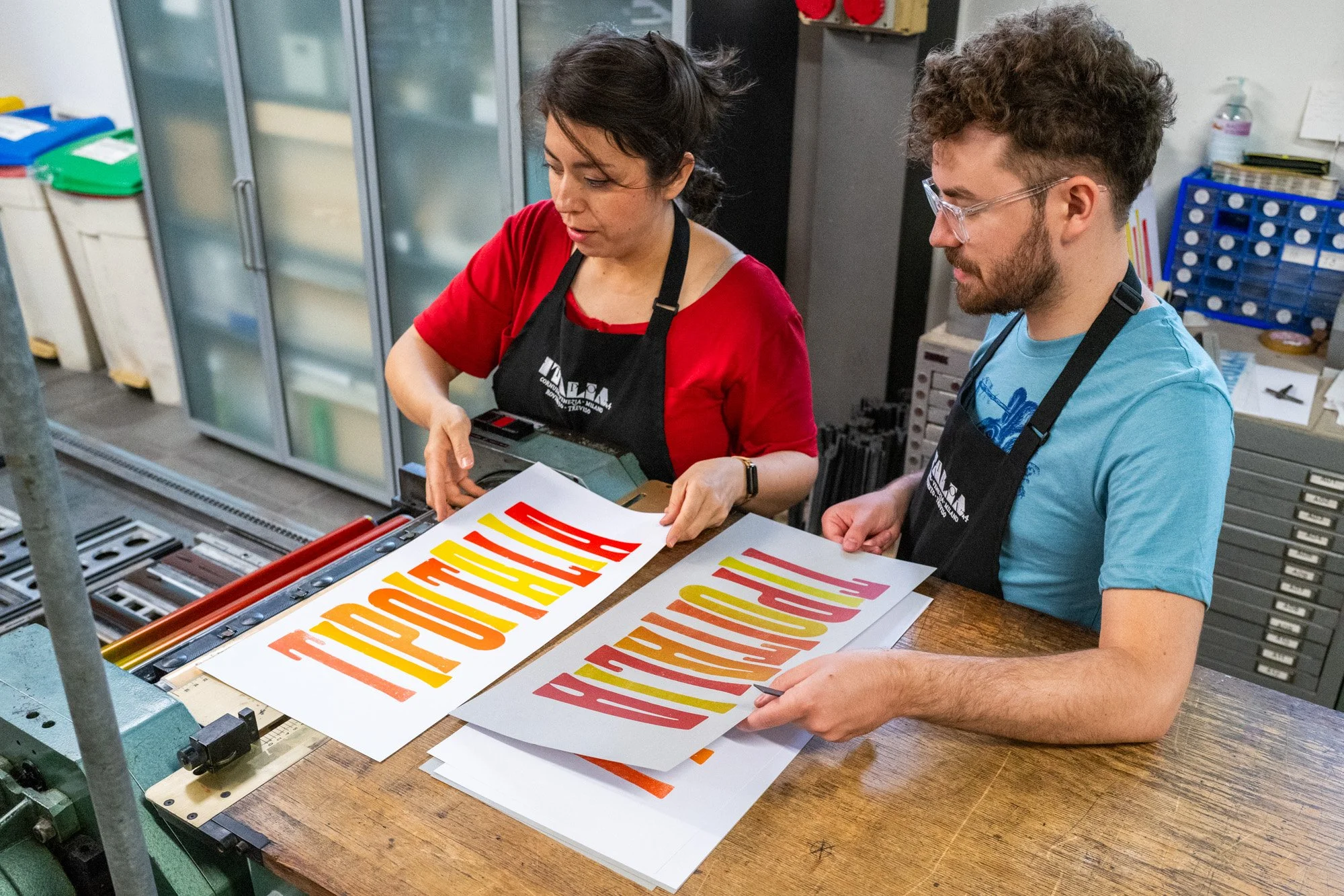Two people are working letterpress prints in the studio at Tipoteca Italiana. The woman on the left is wearing a red top and black apron, and the man on the right is wearing a blue t-shirt and safety glasses.