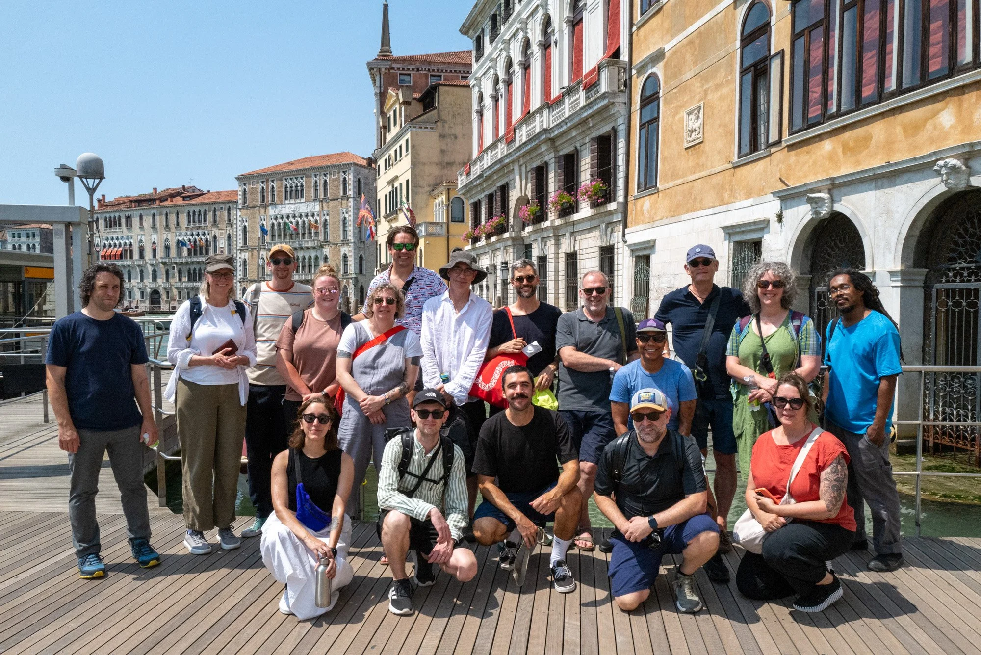 A group of TipoItalia participants posing on a wooden dock with colorful historic buildings and flags in Venice, Italy, in the background.