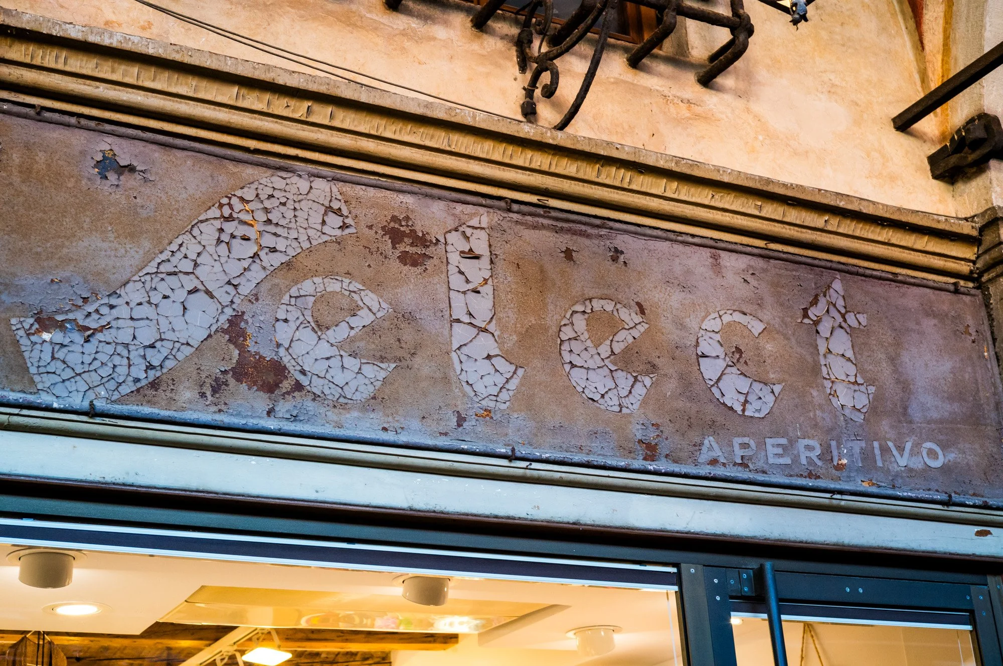 Close-up photo of an old, weathered sign with peeling white paint and cracked letters spelling "Café." The sign appears to be made of metal and has visible rust, located above a storefront entrance.