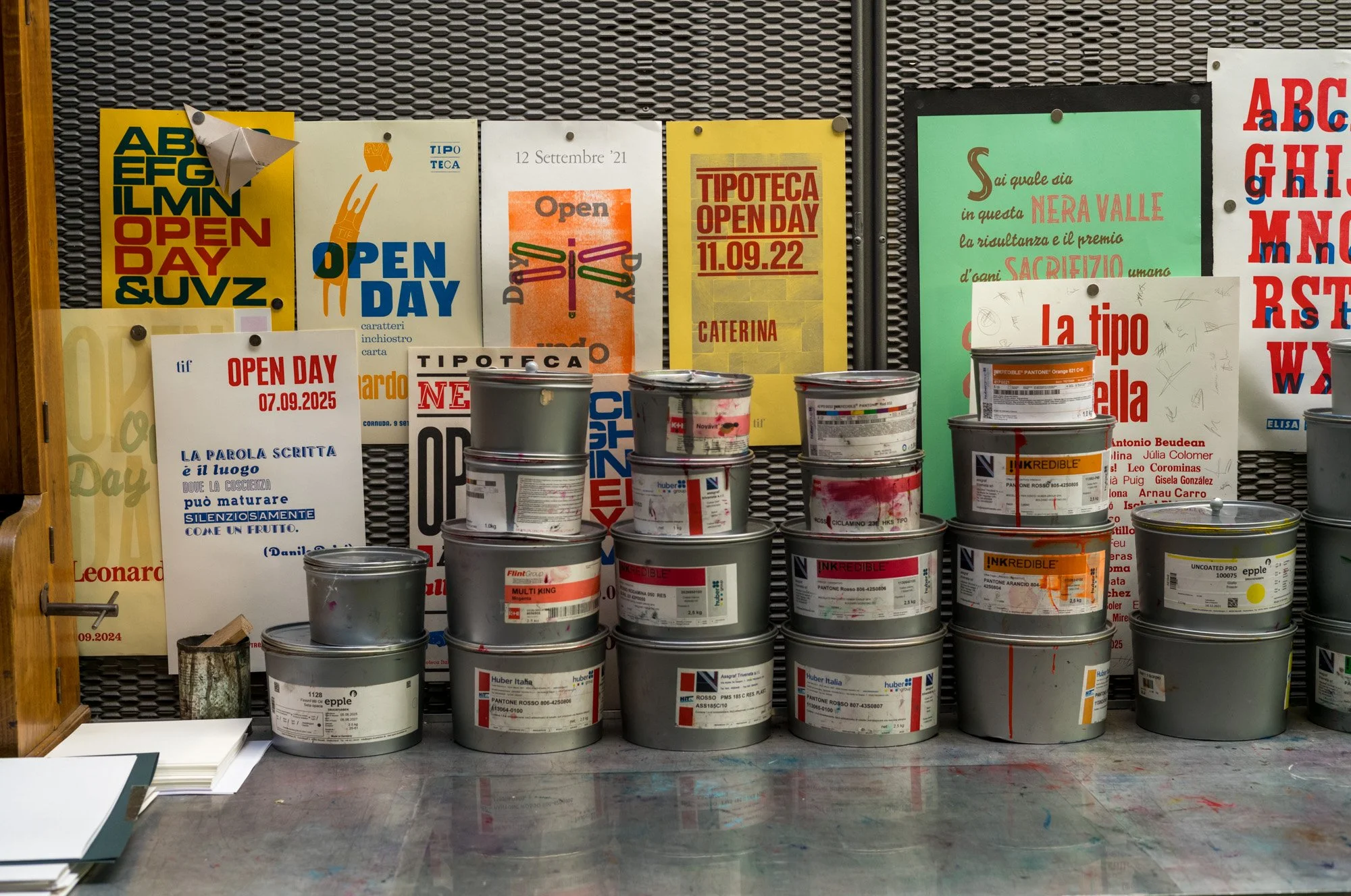 A display of paint cans stacked in front of a wall with colorful posters about open days and events. The table has a few notebooks or papers on it. The posters behind include Italian text and dates.