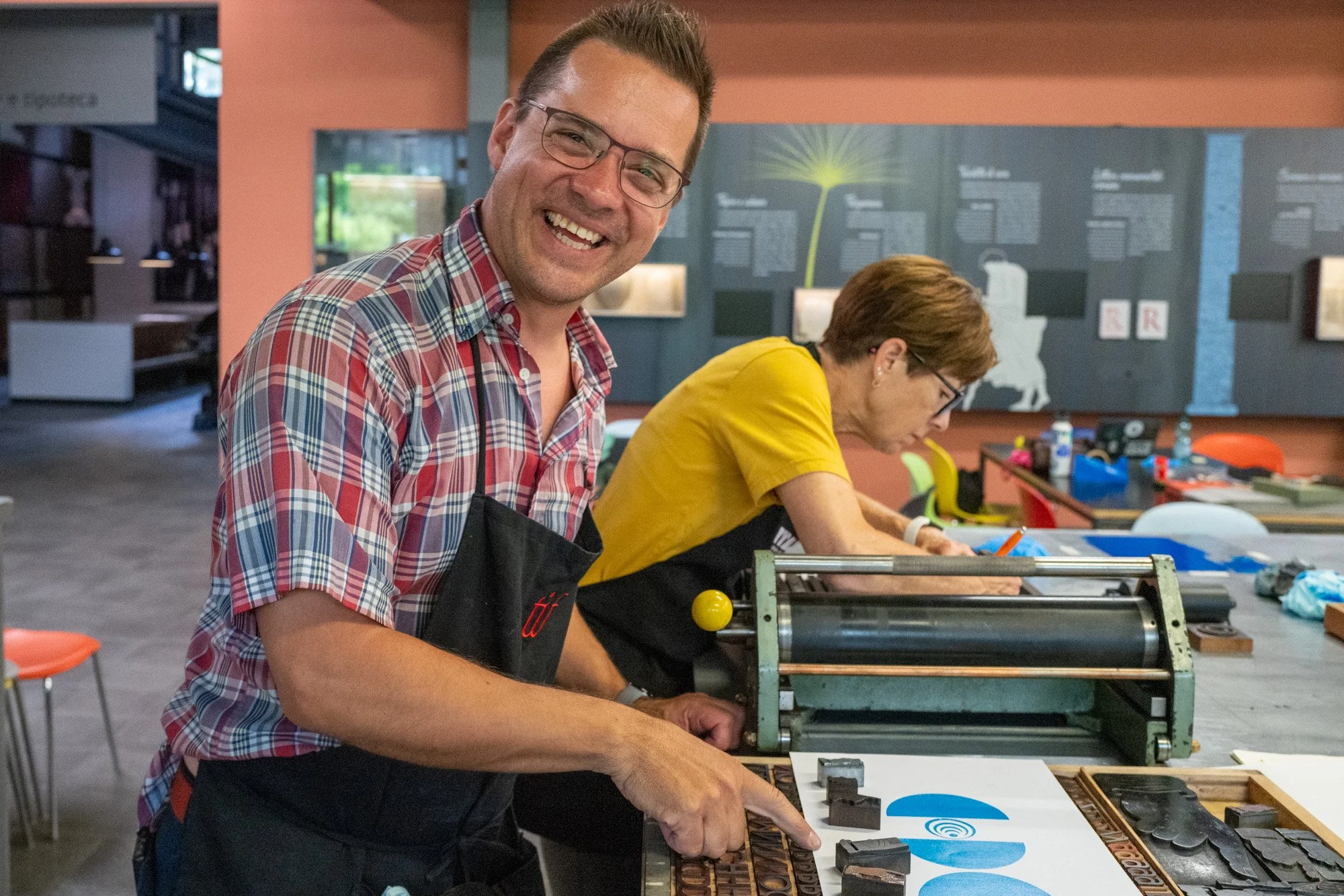 A man smiling while working on a printing press in a creative workshop, with a woman engaged in a similar activity in the background.