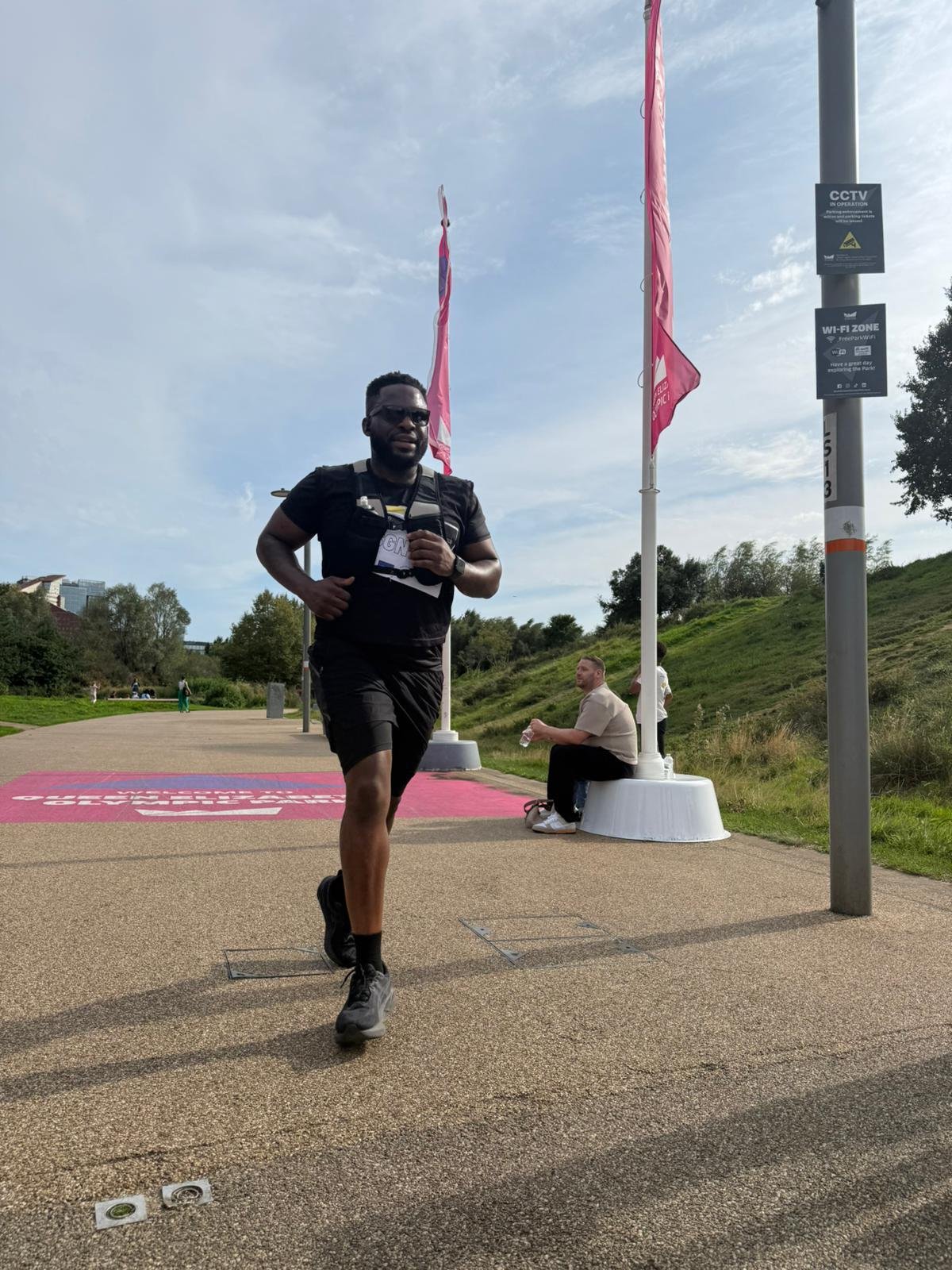 A man running outdoors wearing a black t-shirt, black shorts, gray sneakers, and sunglasses, with flags, a lamppost, and a seated man in the background.