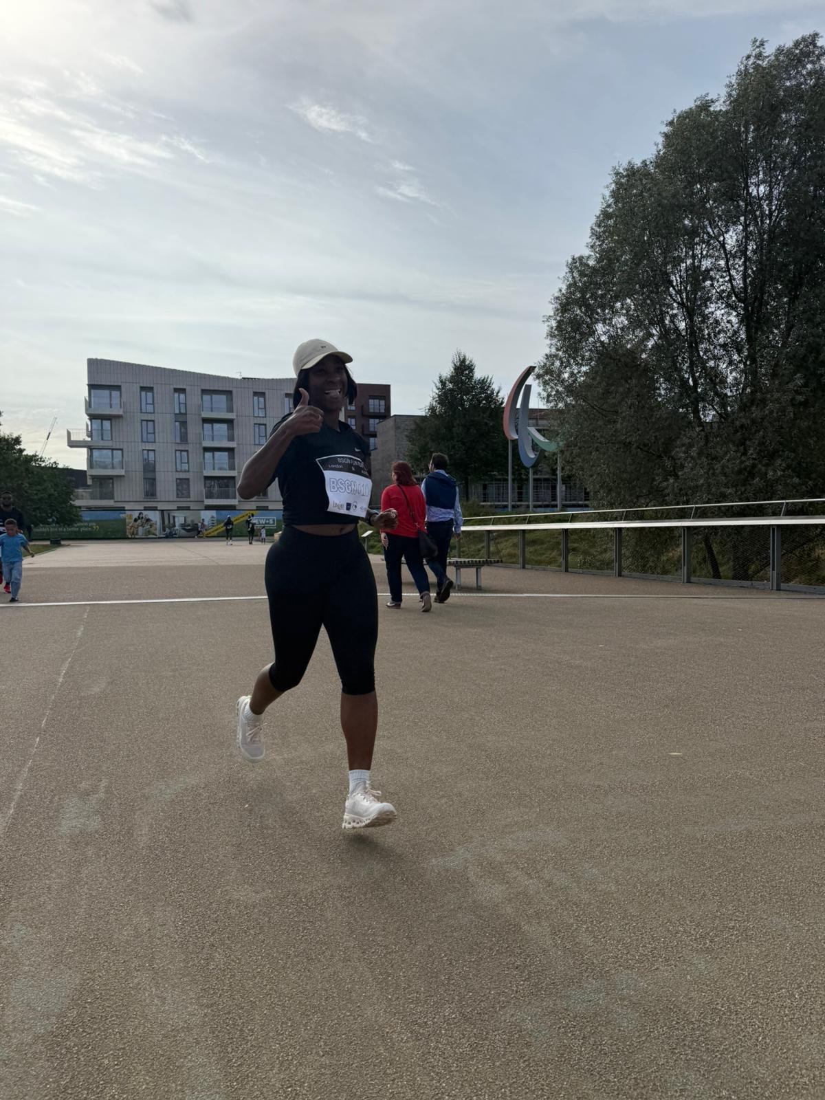 A woman running on a track at a park with buildings and trees in the background, giving a thumbs-up and smiling.