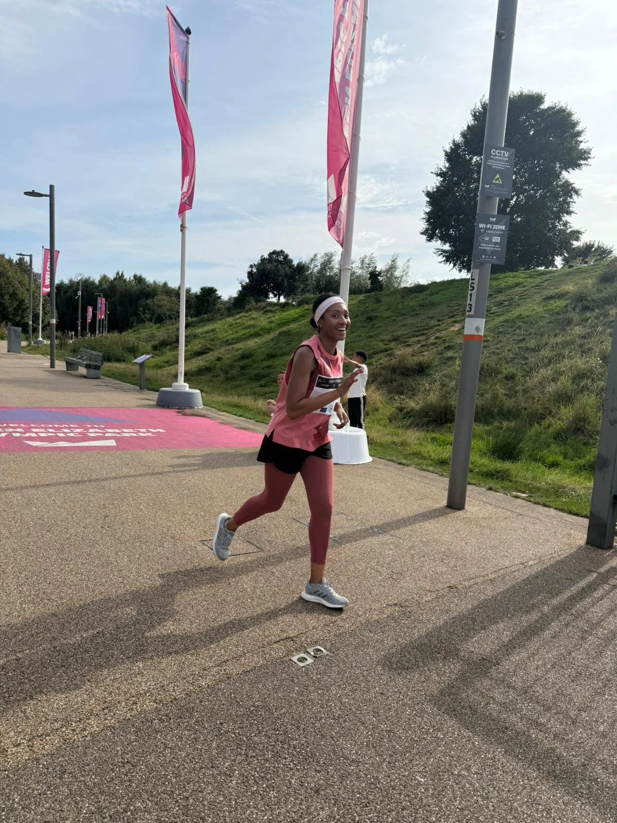 A woman running outdoors during a race, wearing pink athletic gear, a white headband, and smiling, with pink banners and signs in the background.