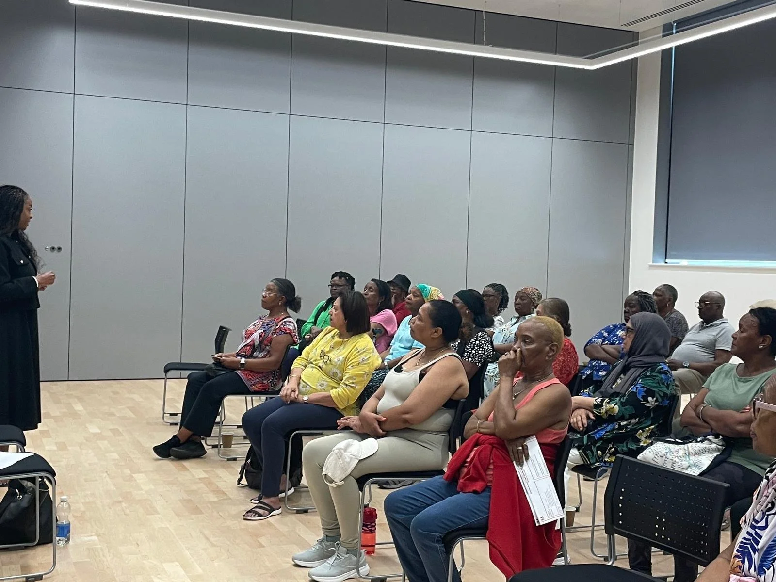 A woman presenter in black attire speaking to a group of diverse women in a modern conference room with gray walls and a black window shade.