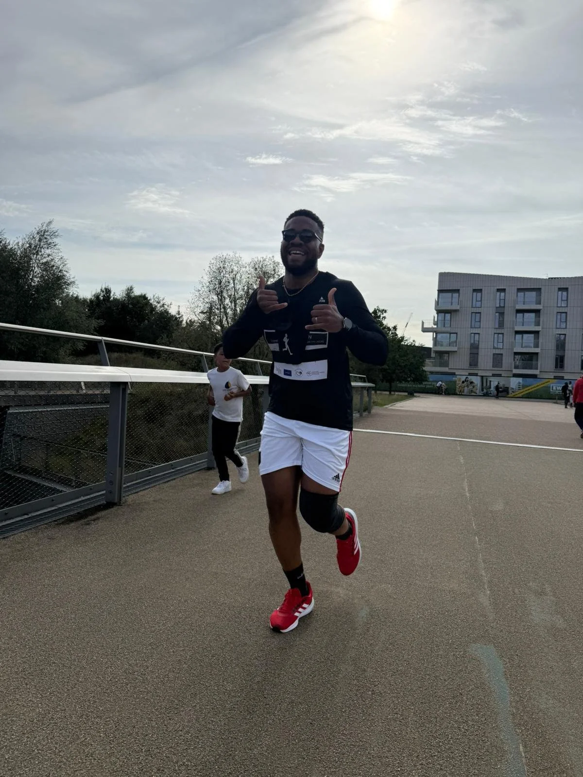 A man running outdoors, giving a thumbs-up and smiling, dressed in athletic gear including red running shoes, white shorts, and a black long-sleeve shirt, with a knee brace on his right knee