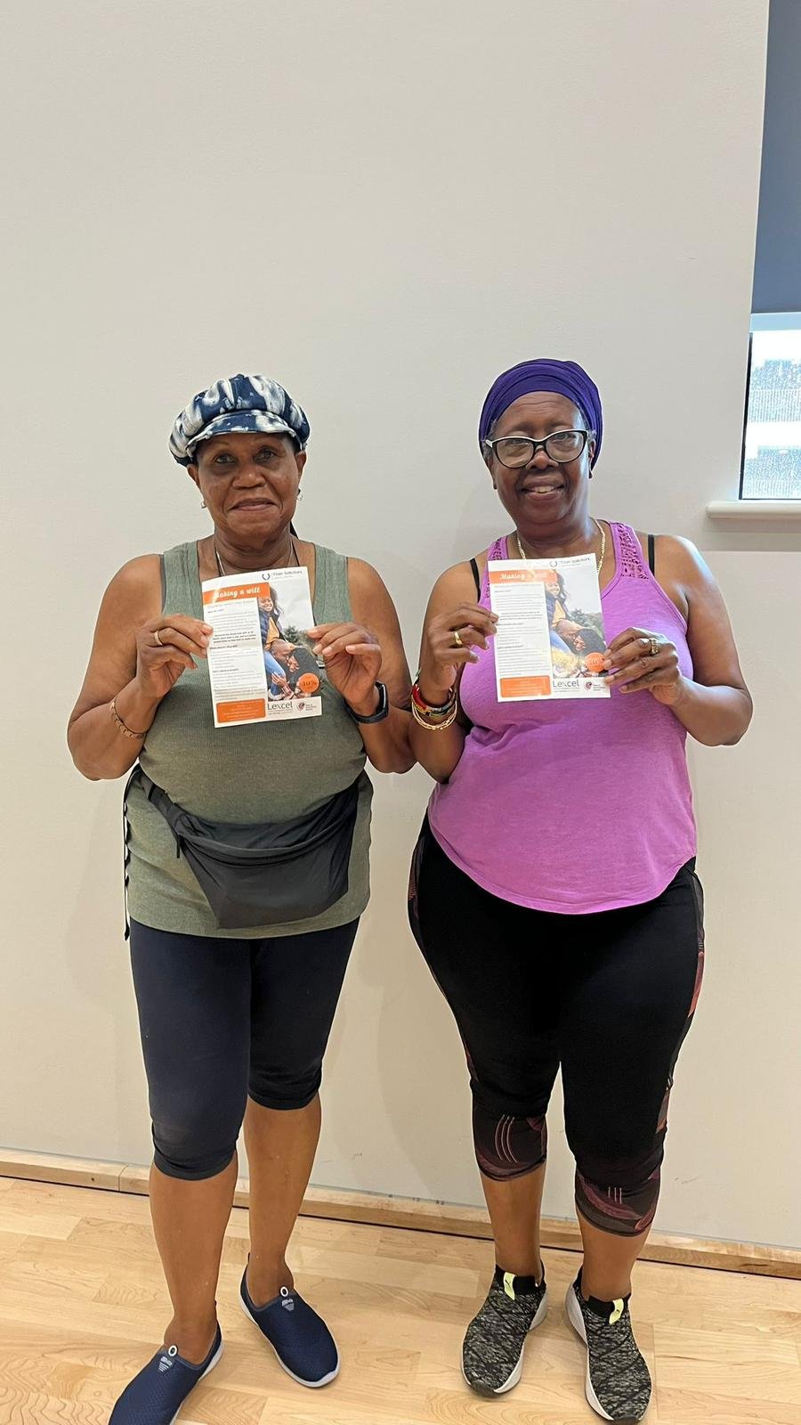 Two women wearing athletic clothes and head coverings, standing indoors against a plain wall, each holding a flyer and smiling at the camera.