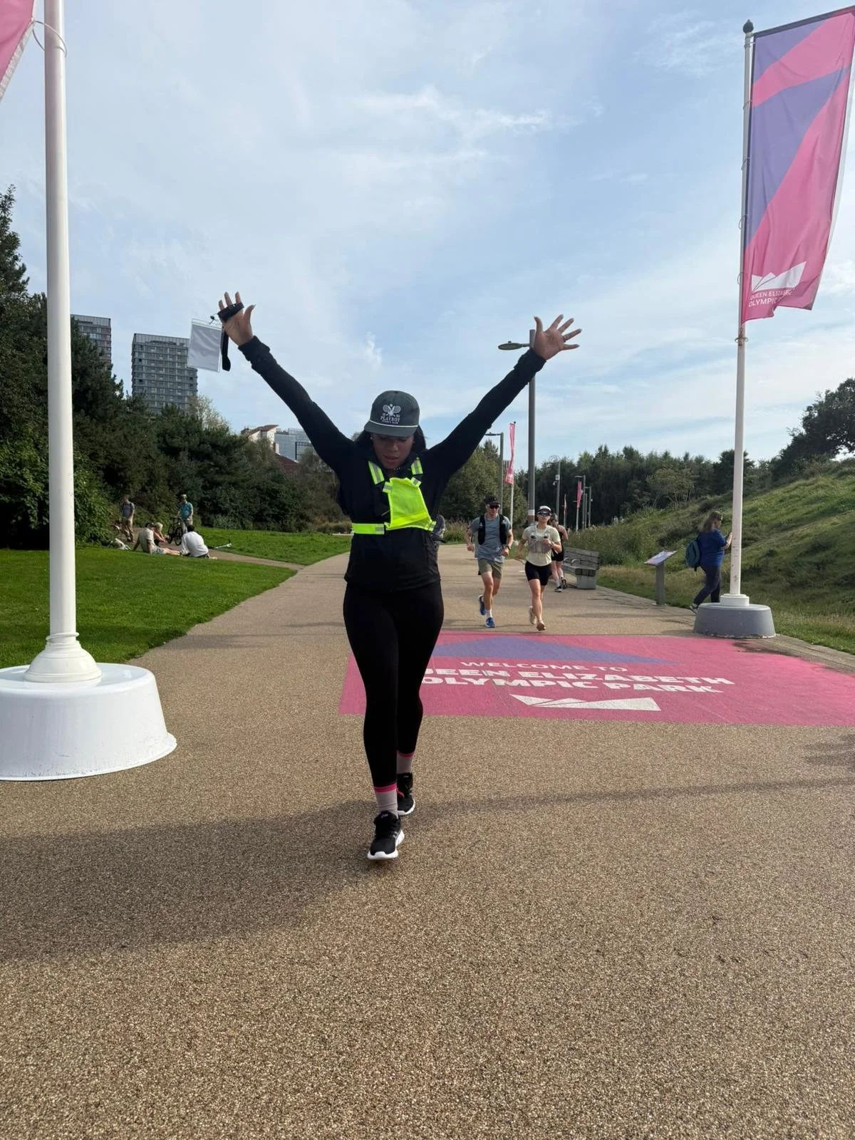 A woman running and celebrating with arms raised during a race or event, with other participants and greenery in the background.