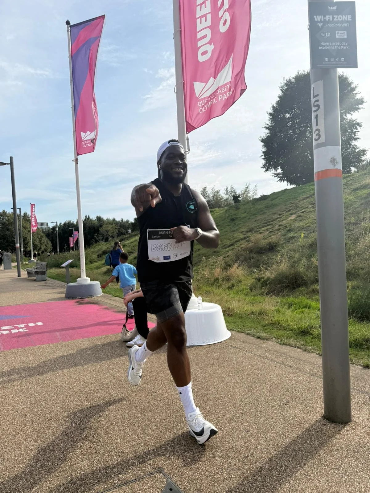 A smiling man running in a race event at Queen Elizabeth Olympic Park, pointing at the camera. He is wearing a black sleeveless shirt, black shorts, white socks, and athletic shoes. There are pink flags and a