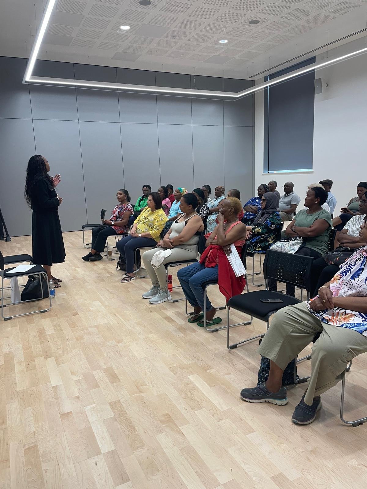 A woman giving a presentation to a diverse group of people seated in a room with modern gray walls, wooden floor, and large window with a blind.