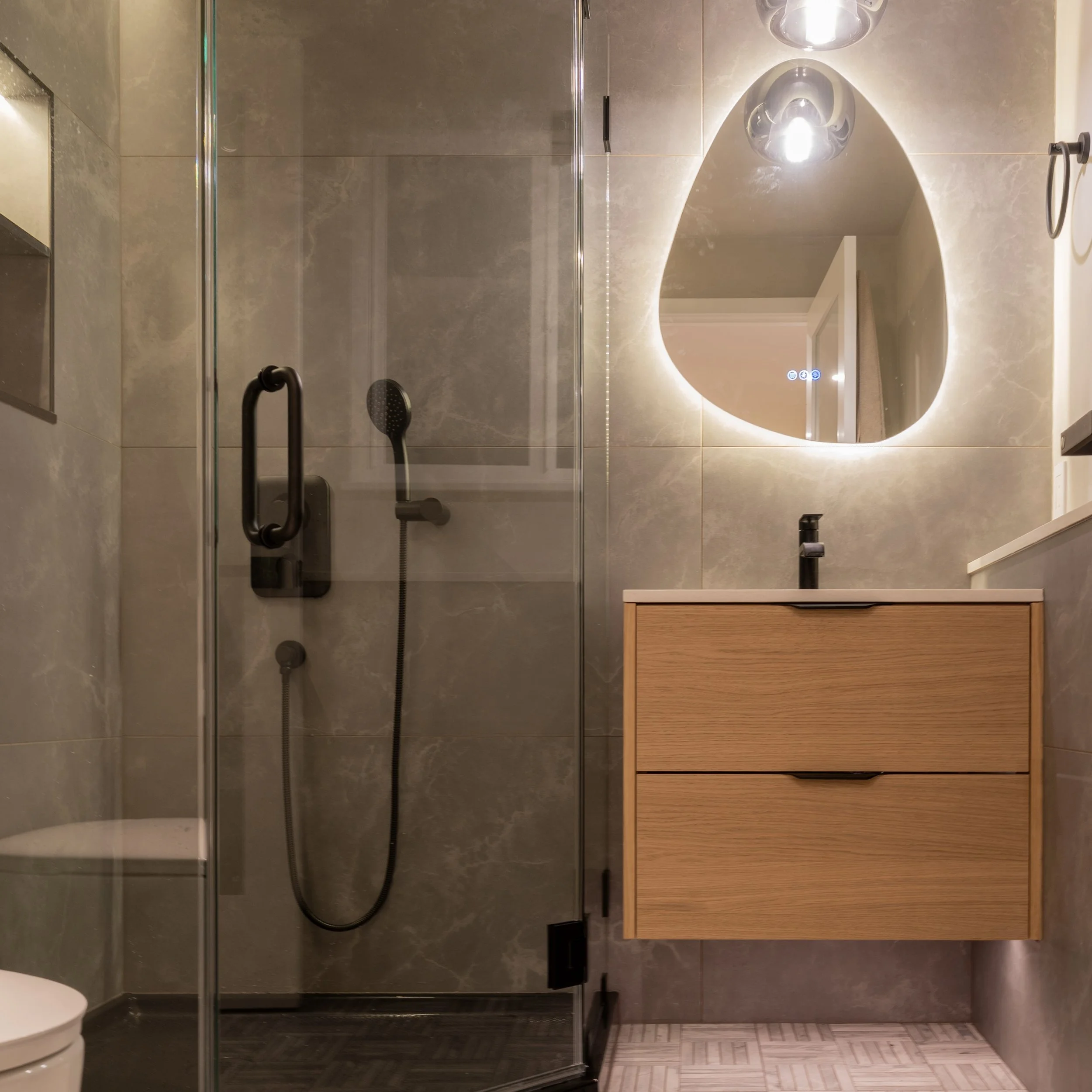 Modern bathroom featuring a glass-enclosed shower with a black handheld showerhead, a wooden vanity with two drawers, and a backlit oval mirror above the sink.