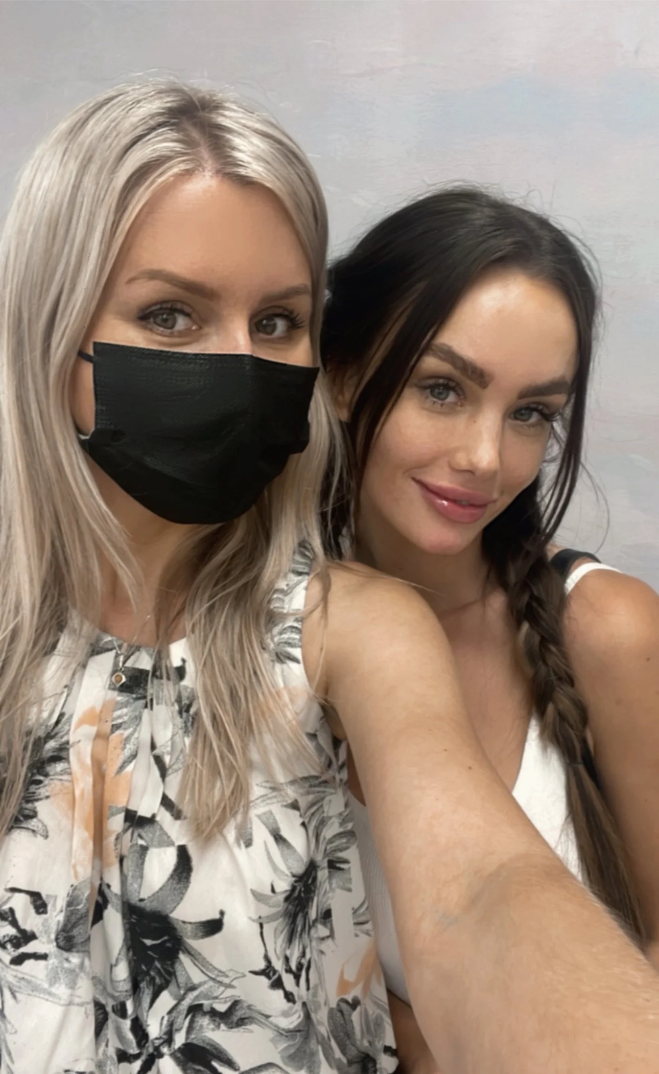 Two women taking a selfie together. The woman on the left has long blonde hair, is wearing a black face mask and a floral top. The woman on the right has long brown hair in a braid, and is wearing a white tank top, smiling at the camera.