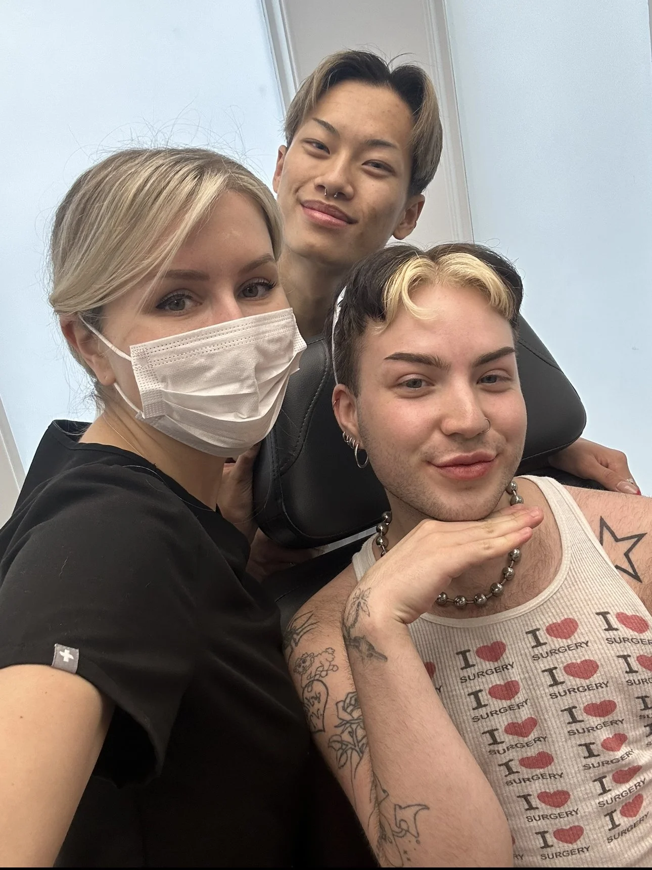 Three people, one wearing a face mask, taking a selfie together indoors. They are close and smiling.