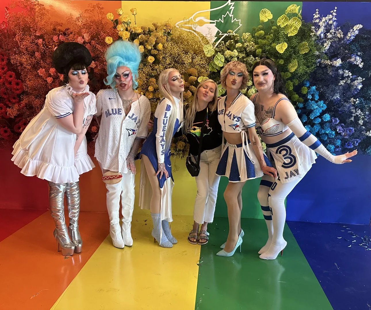 Six people dressed in colorful, team-themed costumes standing in front of a rainbow-colored floral backdrop. The costumes feature the Toronto Blue Jays logo and colors.