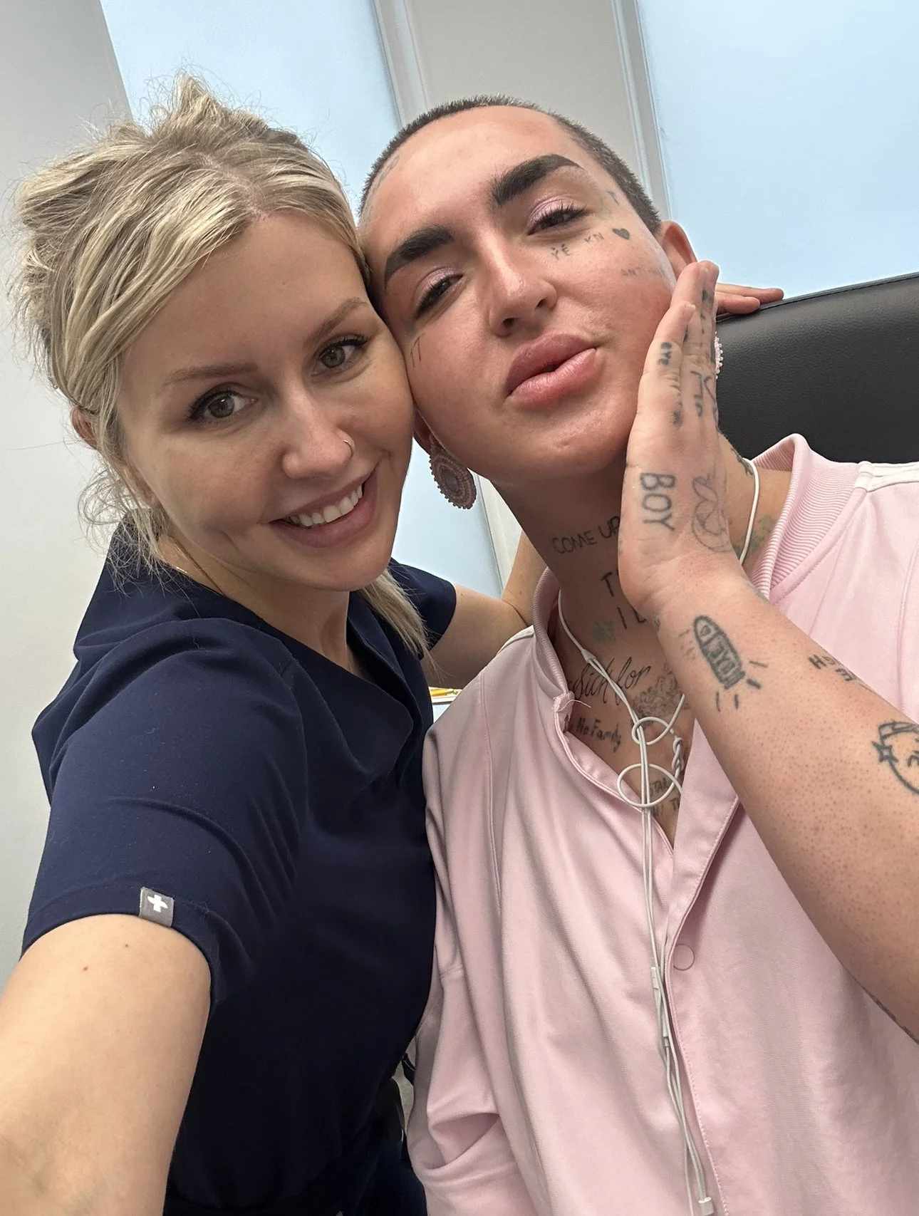 A woman and a man take a selfie together; the woman has light skin, blonde hair, and wears a dark blue shirt, smiling at the camera. The man has light skin, tattoos on his face and arms, and wears a pink shirt, with a relaxed expression. They are in 
