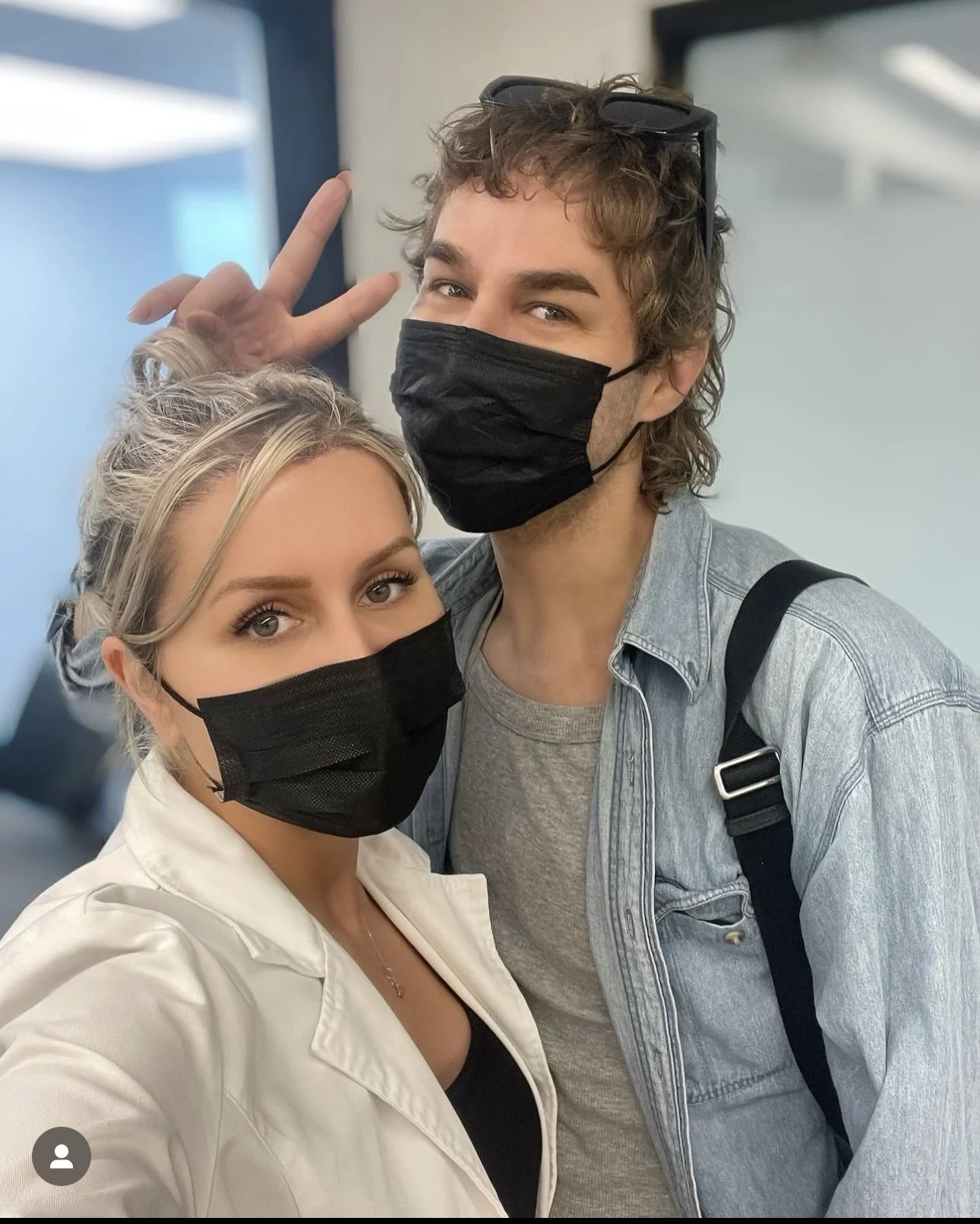 A young man and woman taking a selfie indoors, both wearing black face masks, with the man making a peace sign behind the woman's head, and the woman looking directly at the camera.