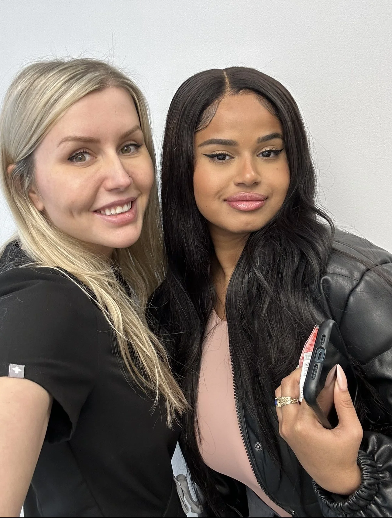 Two women taking a selfie, one with blonde hair and one with dark hair, both smiling slightly, holding a smartphone, standing against a plain white background.