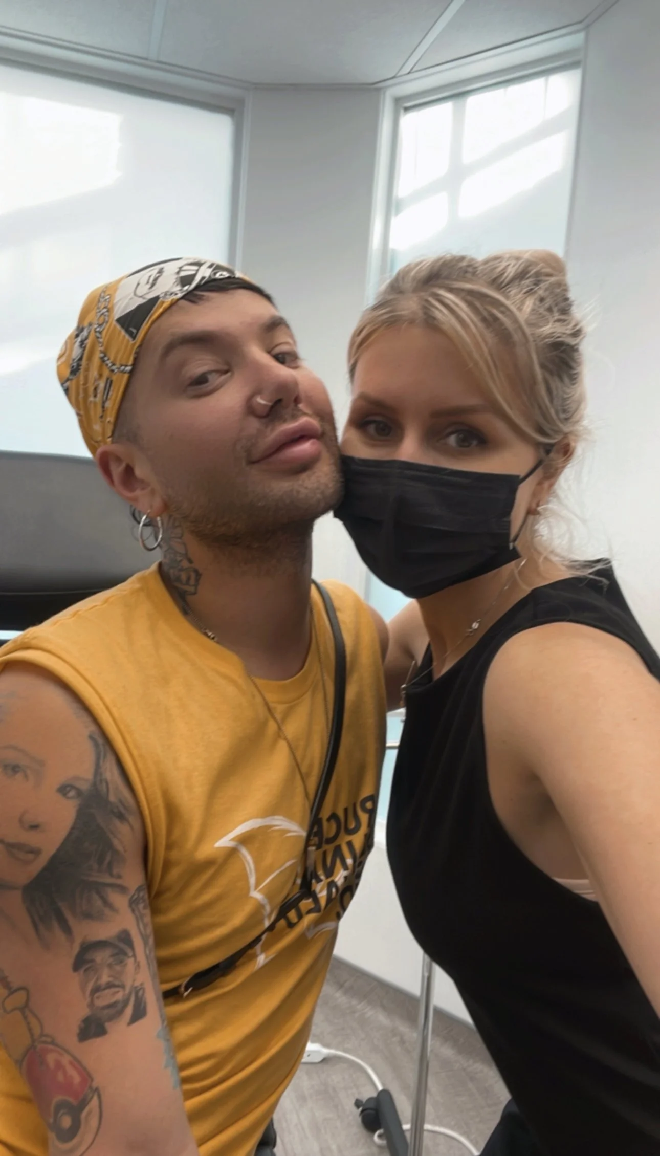 A man wearing a yellow sleeveless shirt, a yellow bandana, and tattoos on his arm is taking a selfie with a woman wearing a black face mask and a black sleeveless shirt, both looking at the camera in a bright room with large windows.