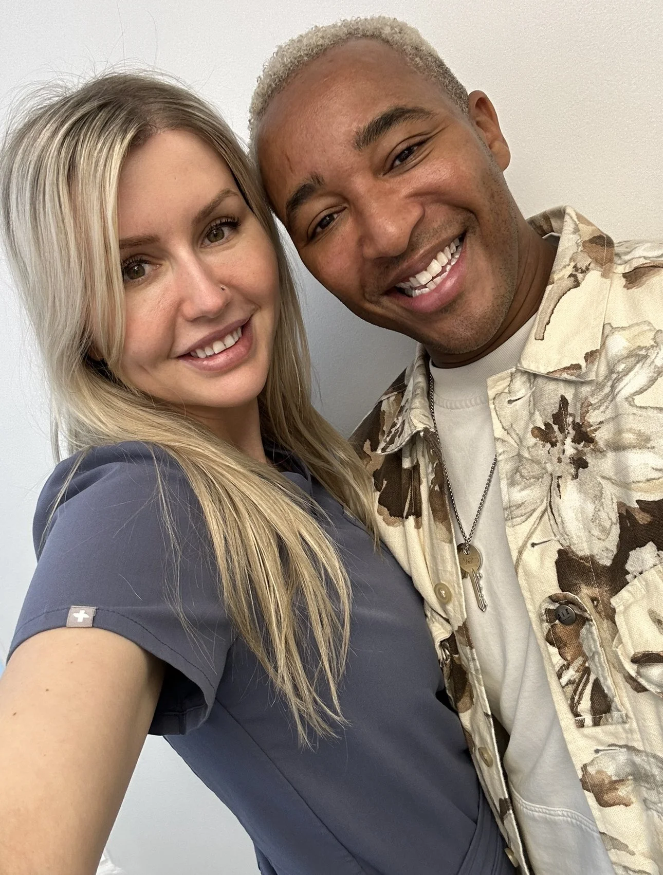A blonde woman in gray scrubs and a smiling man with light blonde hair in a floral jacket take a close-up selfie against a plain background.