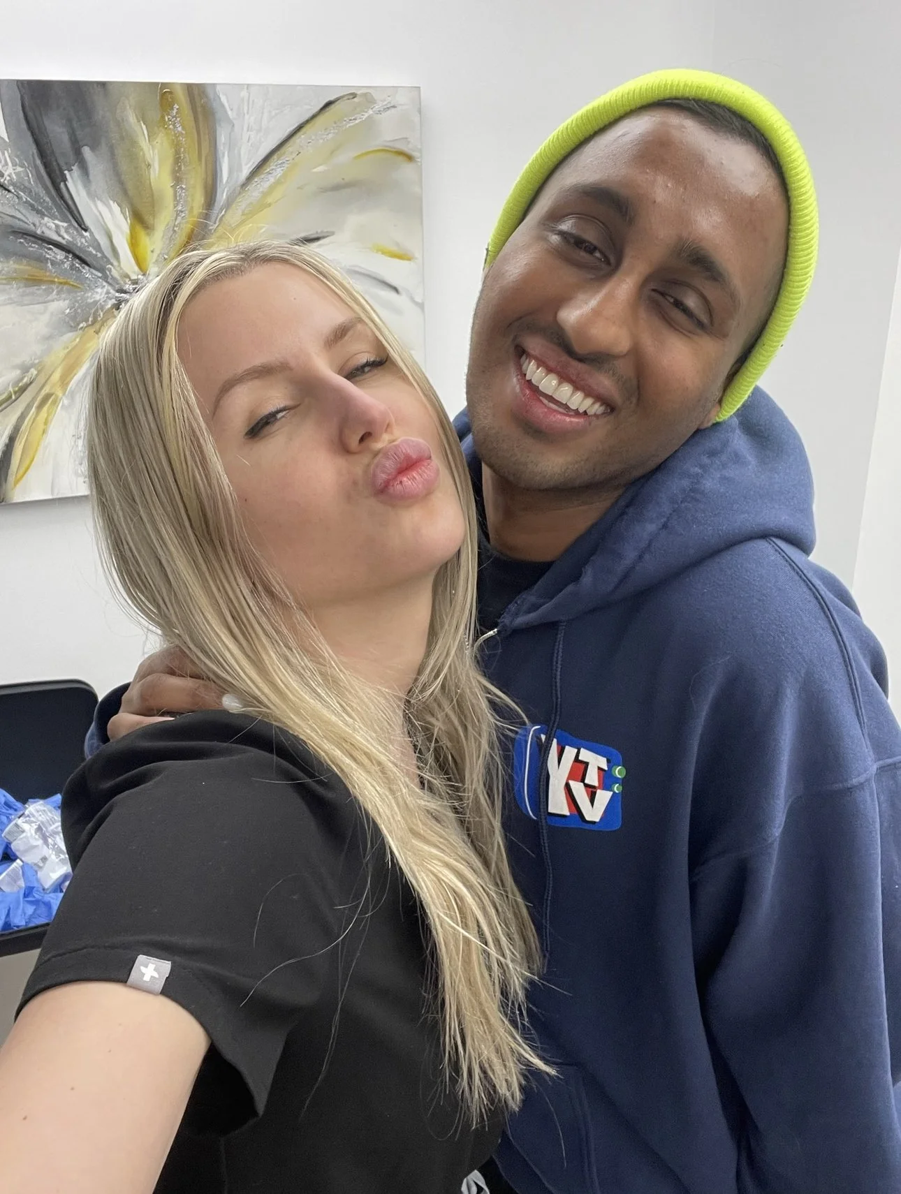A woman and a man taking a selfie together, both making playful facial expressions. The woman has long blonde hair and is puckering her lips, and the man is smiling widely, wearing a bright yellow beanie and a blue hoodie with a logo.