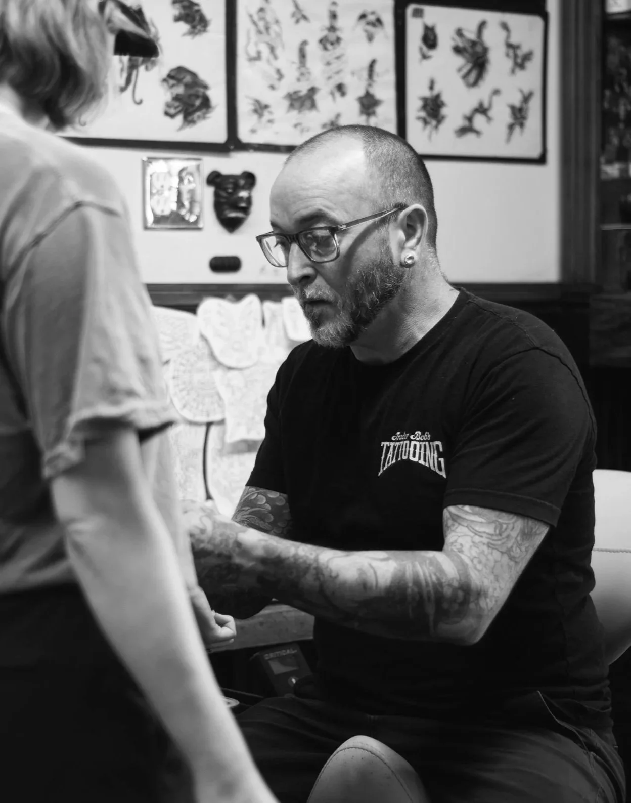 A tattoo artist with tattoos on his arms, glasses, and a beard, giving a tattoo to a customer in a tattoo shop, with various tattoo designs displayed on the wall in the background.