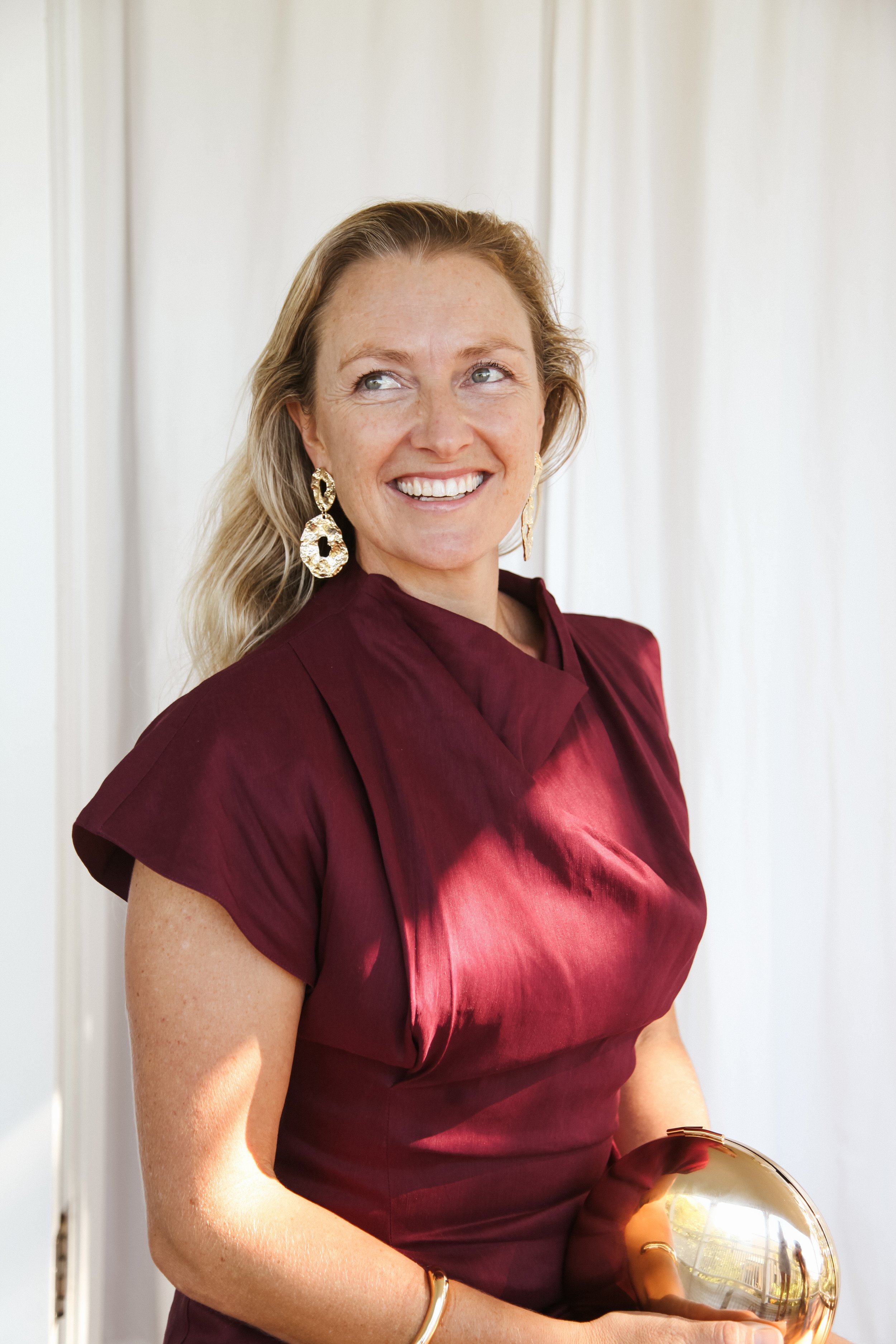 A smiling Skye Swift with long blonde hair wearing a burgundy dress and gold earrings, holding a shiny gold clutch purse, standing in front of a light-colored background.