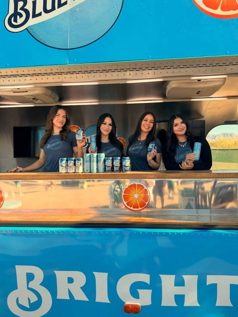 Four women standing behind a promotional juice stand, holding cans of juice, with cans displayed in front of them and a large blue sign with the word 'Bright' and an orange slice symbol.