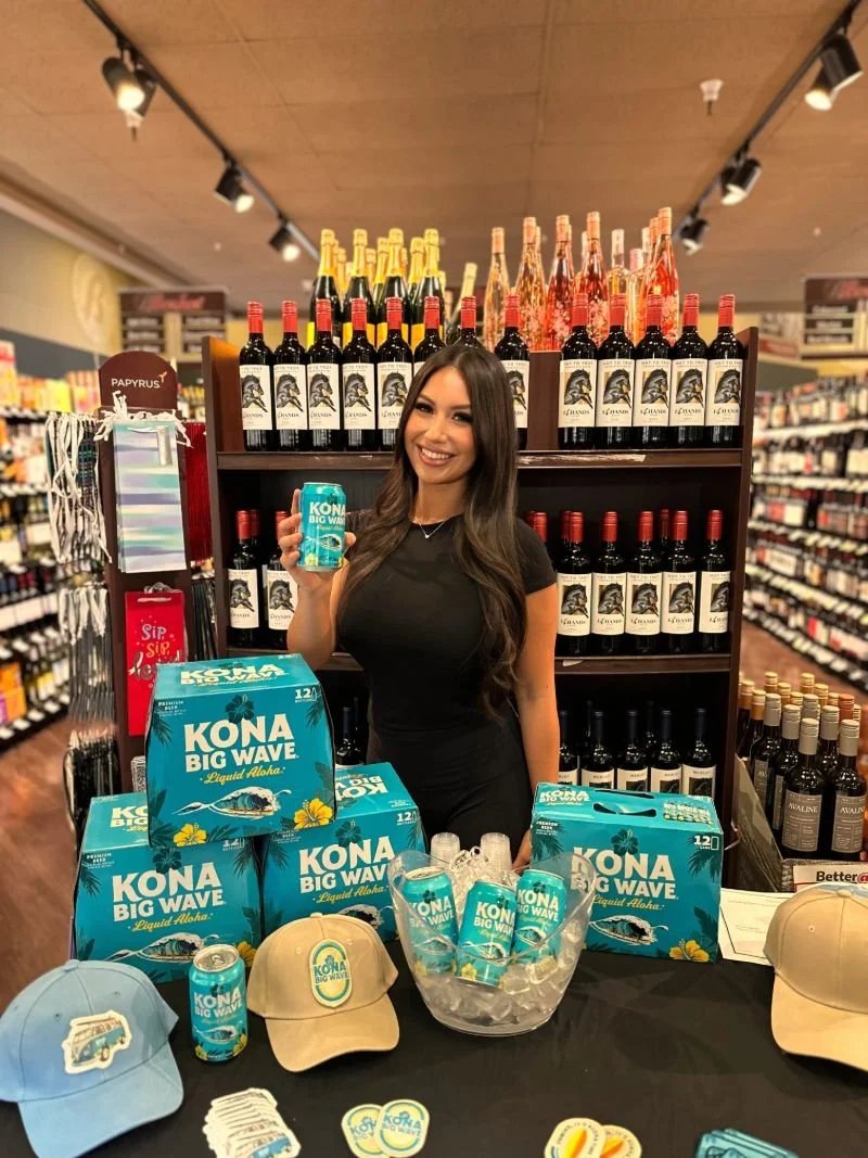 One of EPI's female Brand Ambassadors stands at a promotional booth for Kona Big Wave beer, holding a can with shelves of wine bottles in the background at an on-premise event at a local liquor store to provide premium sampling of Kona Big Wave beer