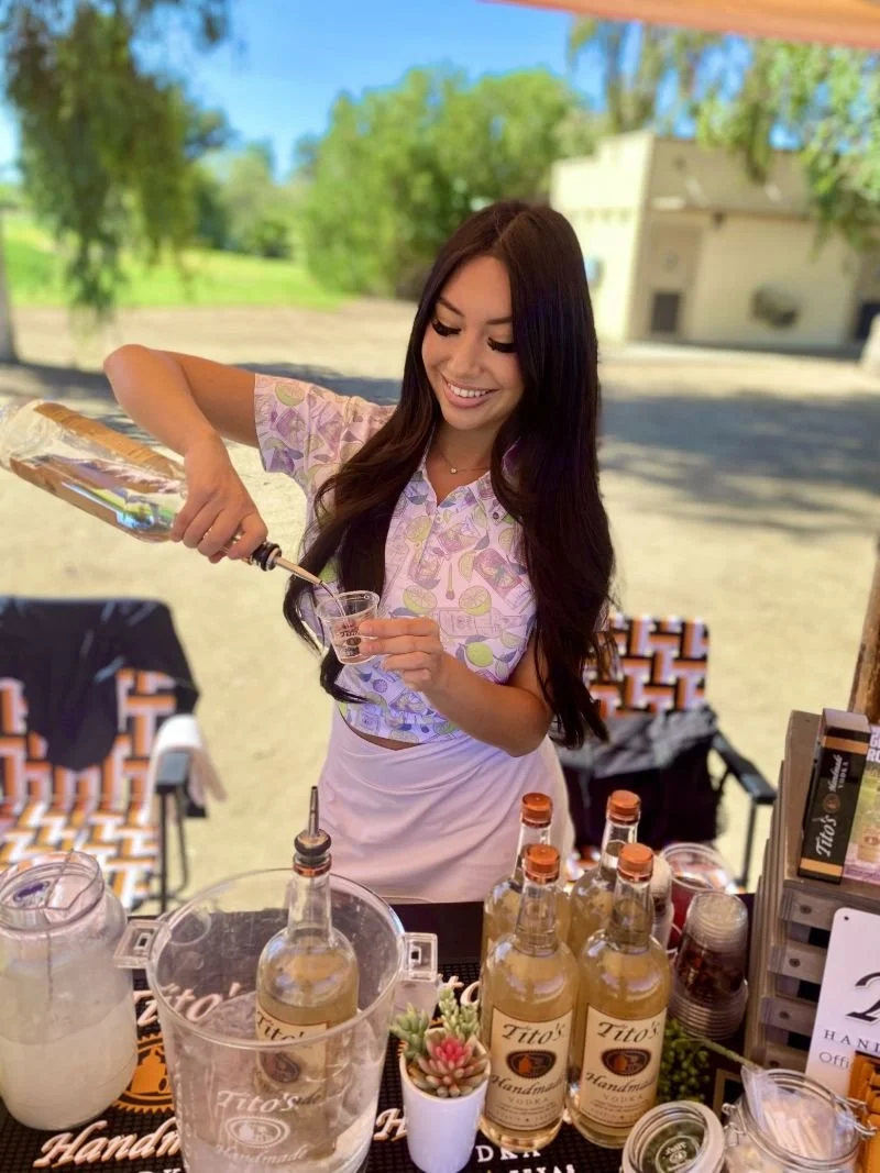 A female Brand Ambassador with long dark hair pouring Tito's Handmade vodka into a sampling cup at an outdoor activation, with bottles of Tito's Handmade Vodka on the table in front of her.