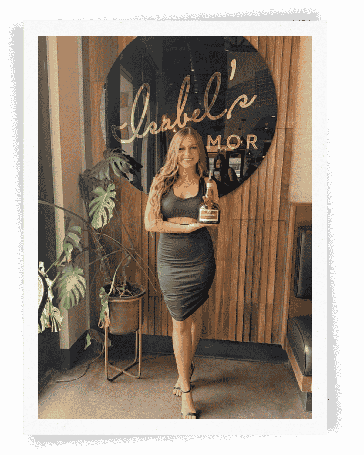 One of our Brand Ambassadors in a black dress with high heels, holds a bottle of Grand Marnier in front of a wooden wall with a round sign that reads 'Isabel; AMOR' -- a Tequila Bar in Gilbert, AZ -- for a high-end, private sampling event