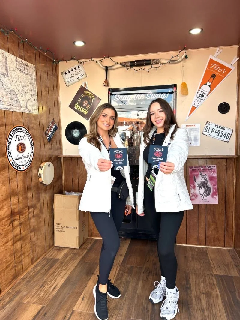 Two EPI Promotions brand ambassadors in white jackets holding Tito's Handmade Vodka coozies inside a branded wood-paneled on-premise activation, representing premium beverage staffing by a national brand ambassador agency.