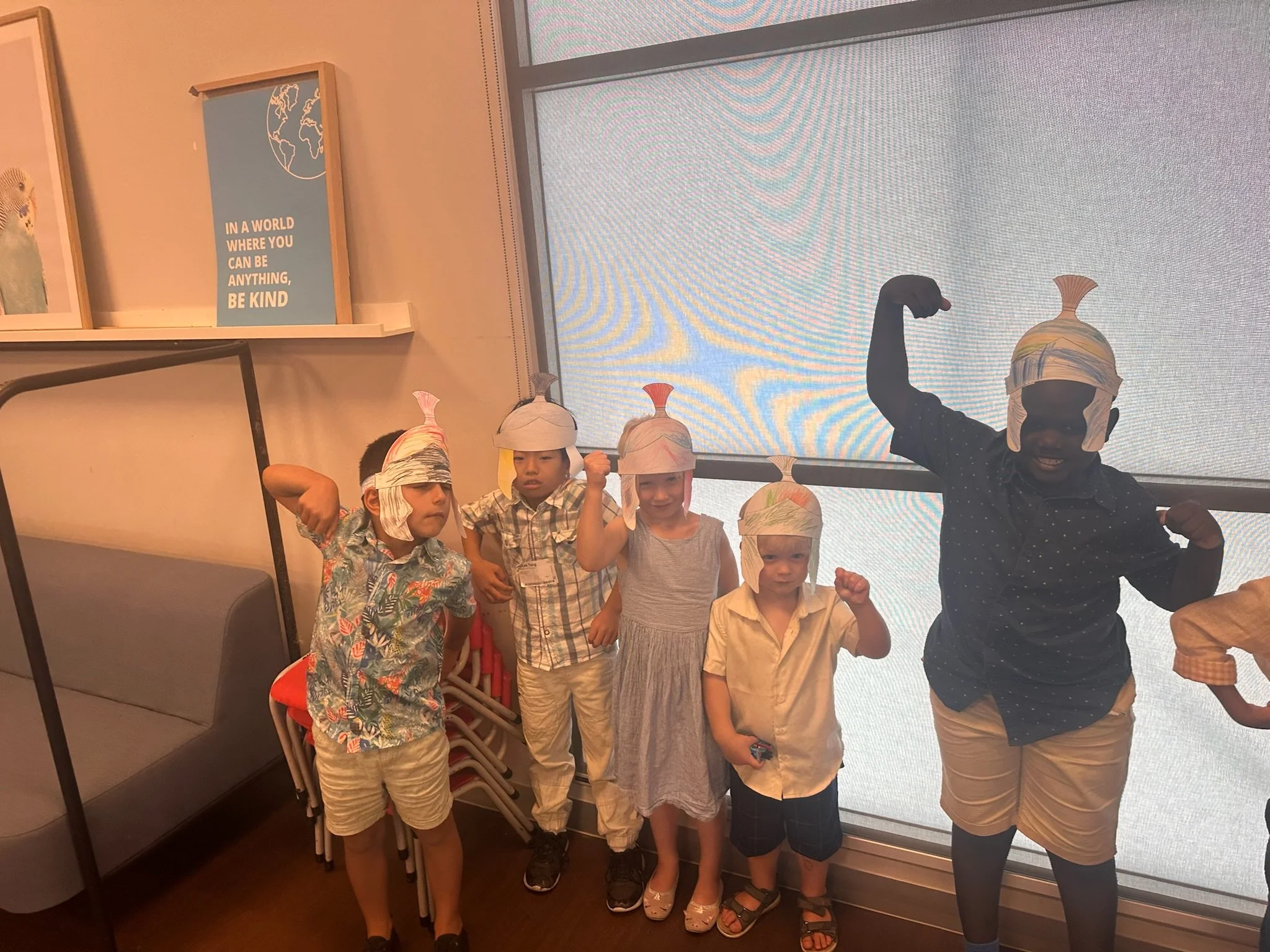 Children wearing paper fish hats and flexing their muscles indoors near a large window and a wall with framed art and a motivational quote.
