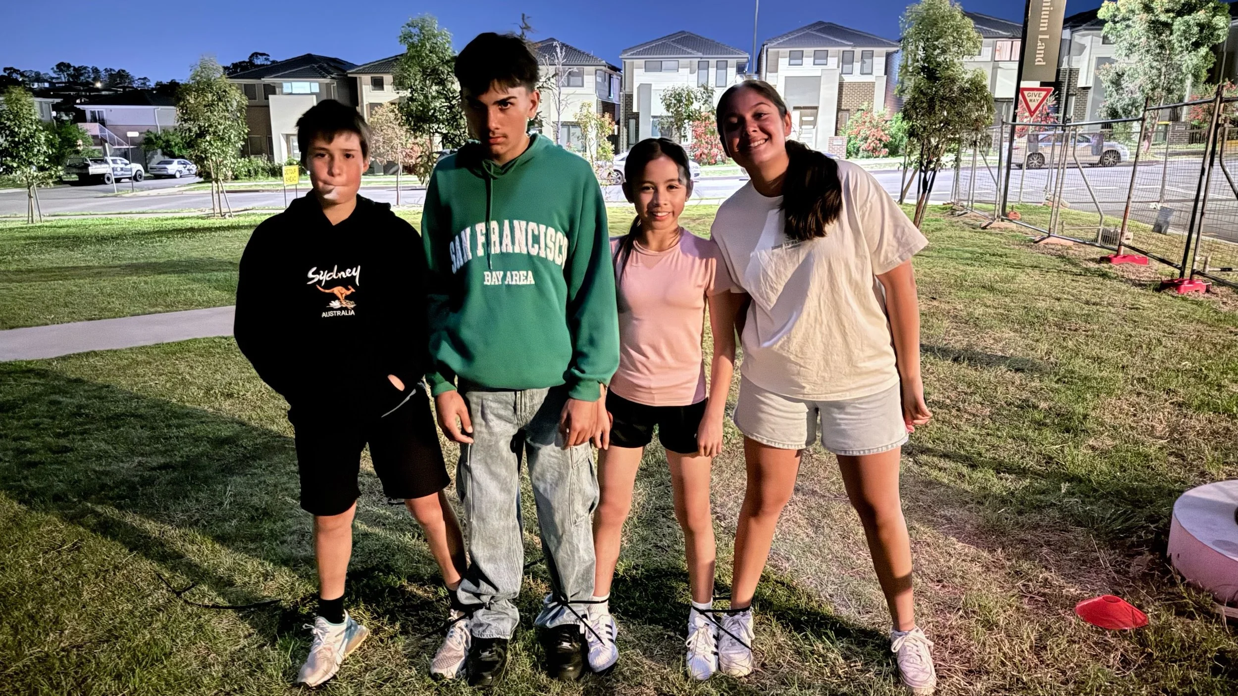 Four teenagers standing on a grassy field at dusk, with residential houses and trees in the background. They are posing for a photo, dressed casually.
