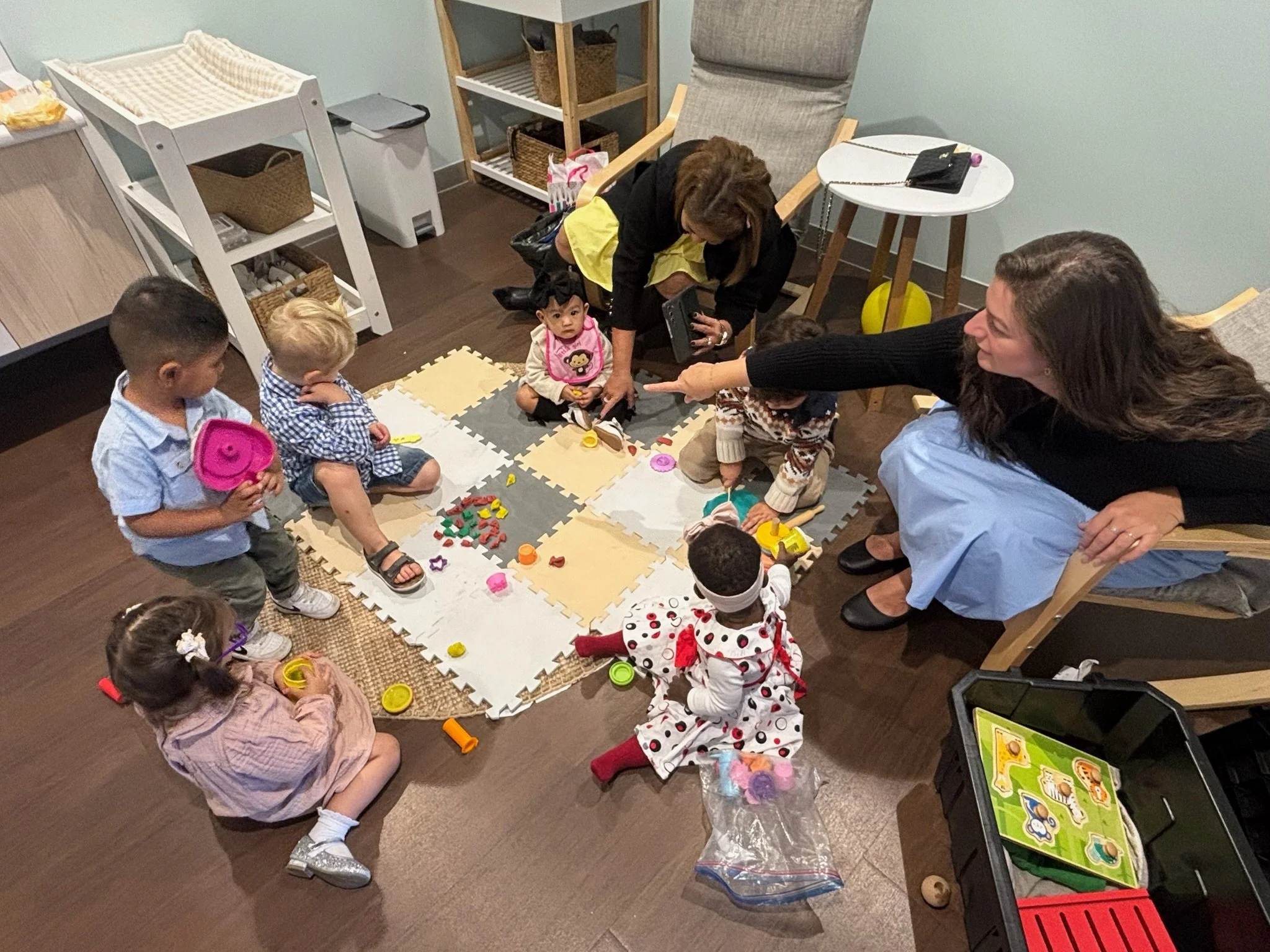 Children playing with toys on foam puzzle mats while two women interact with them in a playroom.