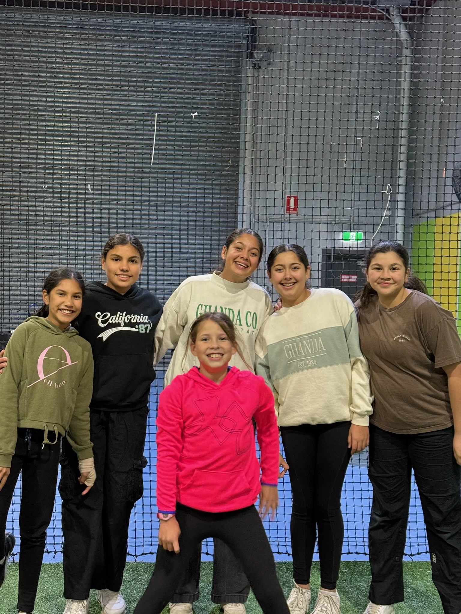 Group of six young girls smiling and posing indoors in front of a sports net and blue court.