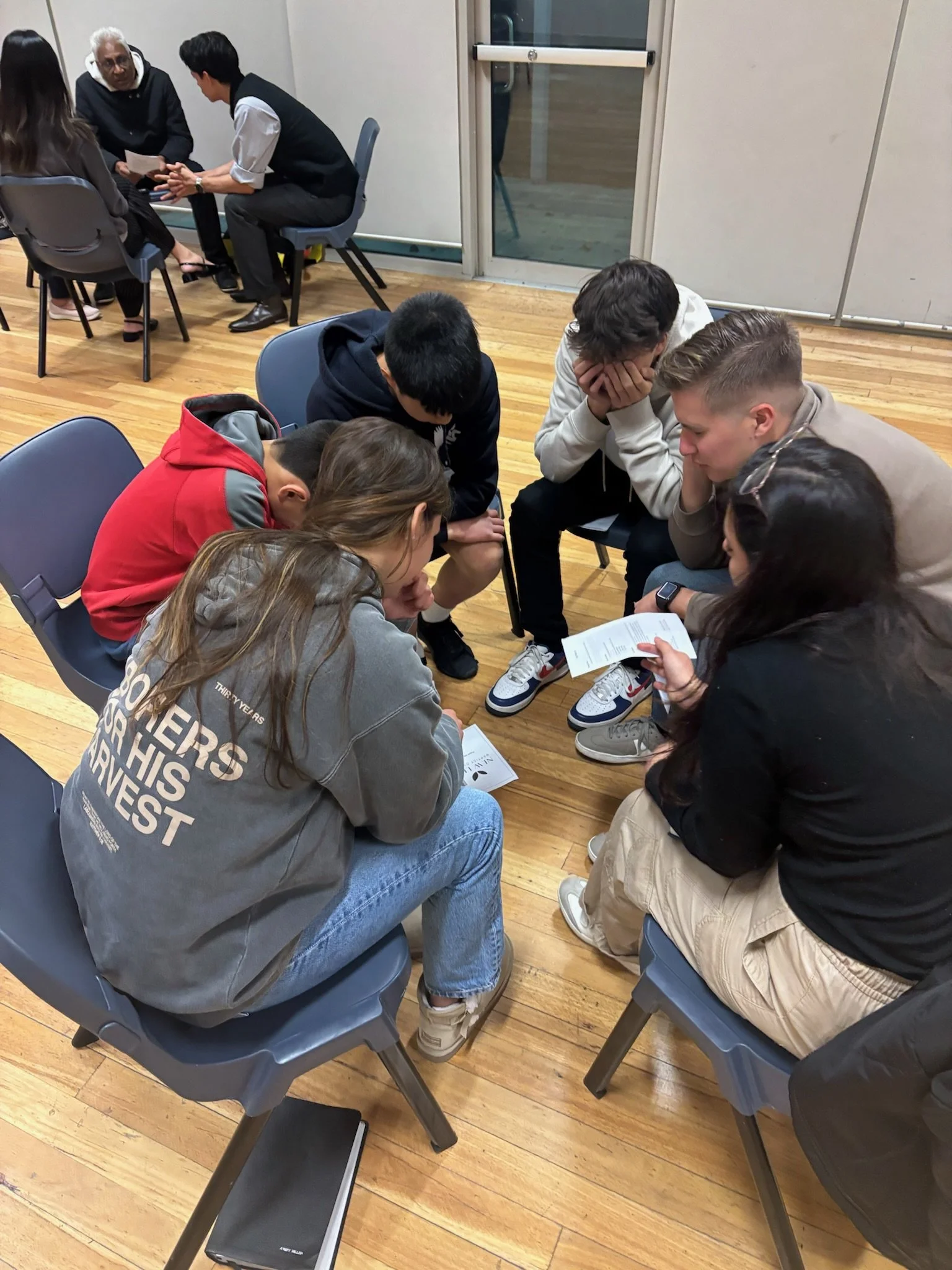 Group of young people sitting in a circle on chairs, engrossed in a discussion or activity, with two more individuals at a small table in the background.