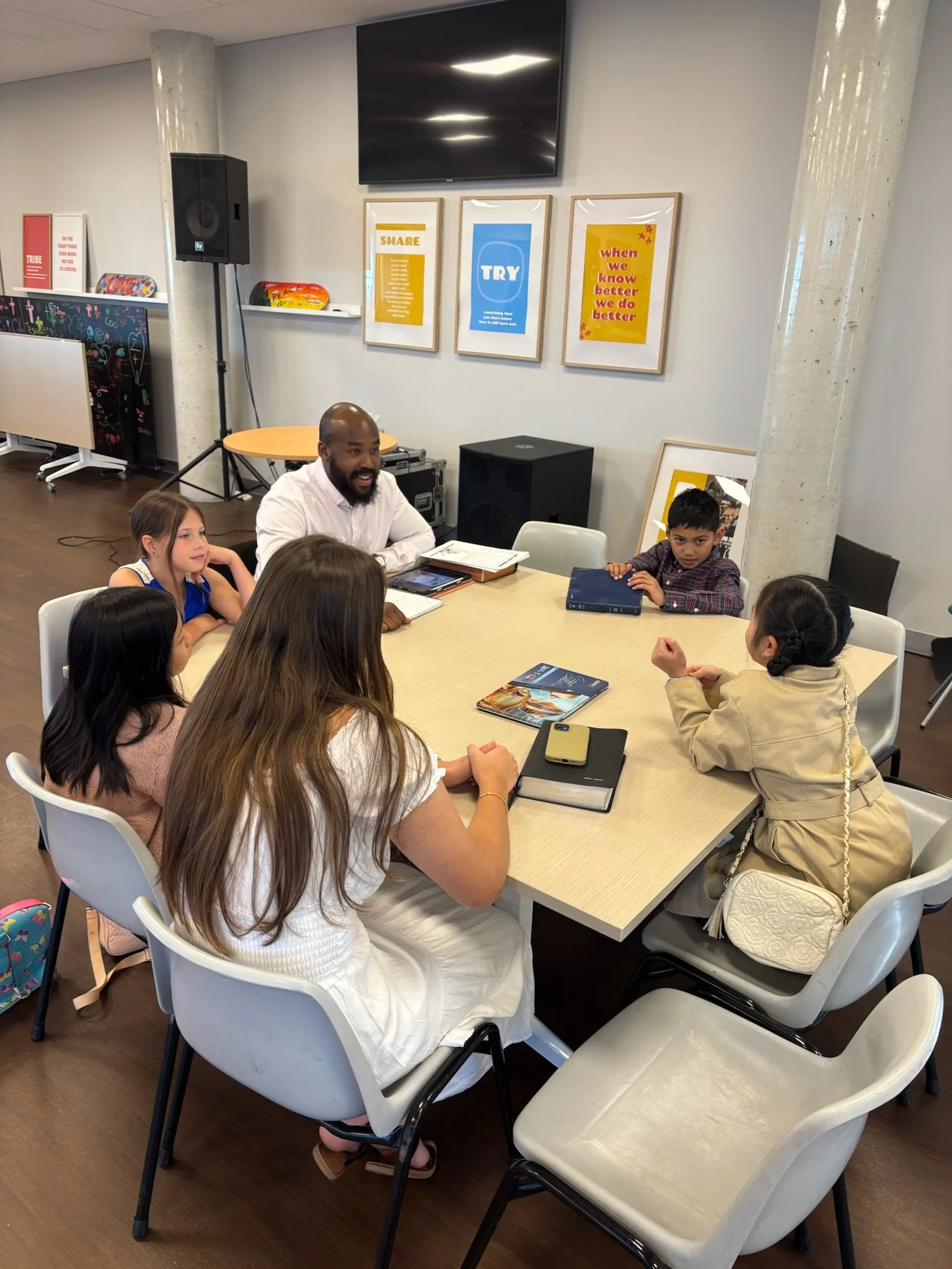 A group of five children and a man are sitting around a table in a classroom or meeting room, engaged in conversation or learning. There are several books and notebooks on the table, and posters on the wall behind them.