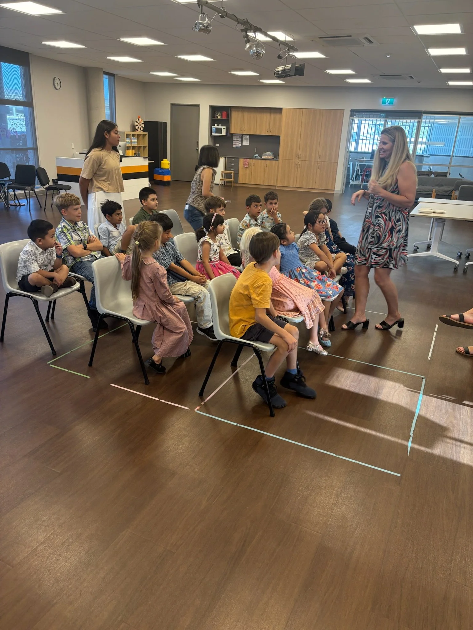 A group of children sitting on chairs in a classroom or community room, with a woman standing in front of them speaking or teaching and two women standing behind the children.