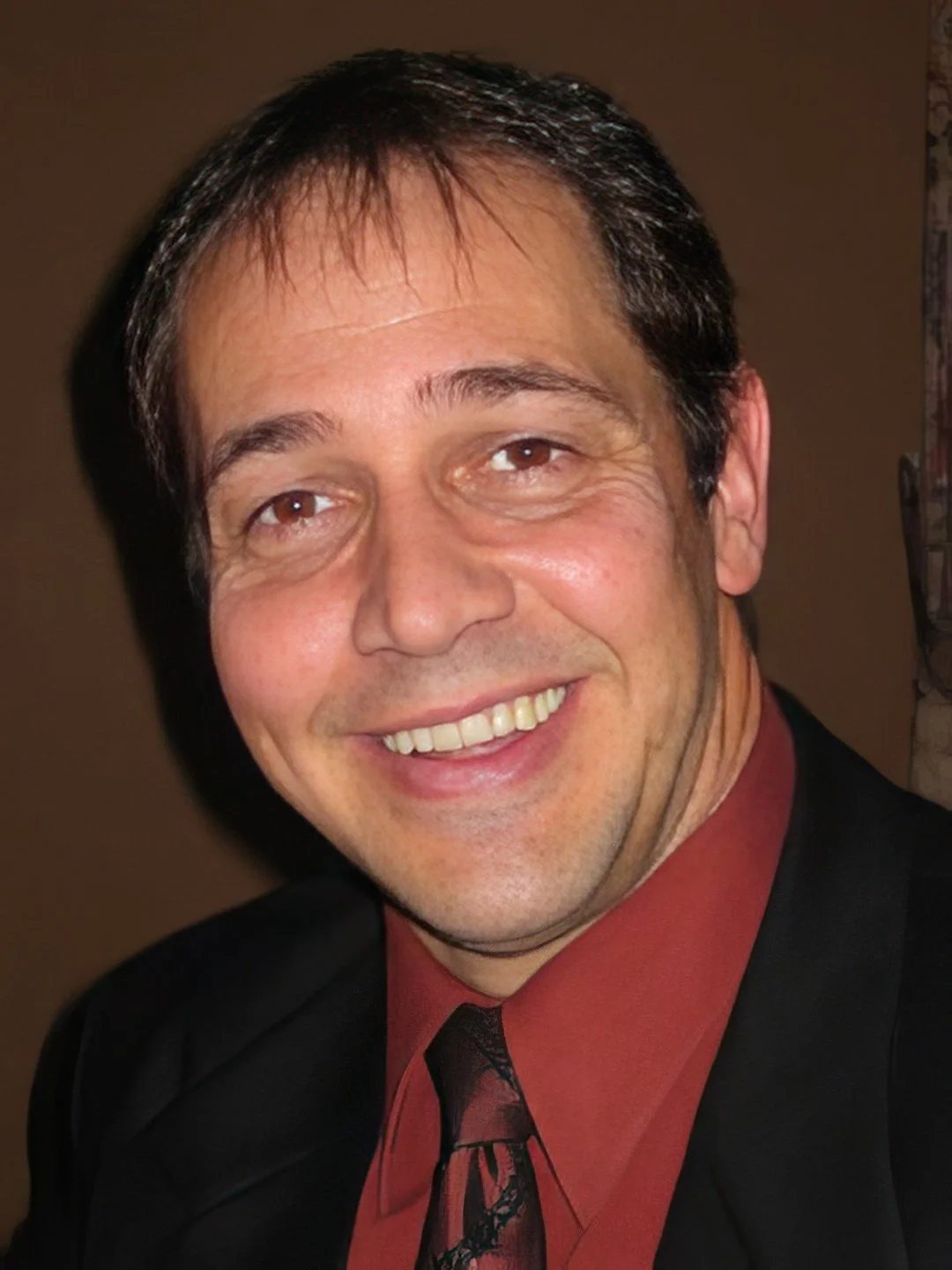 A smiling man with dark hair wearing a black suit jacket, red shirt, and patterned tie.