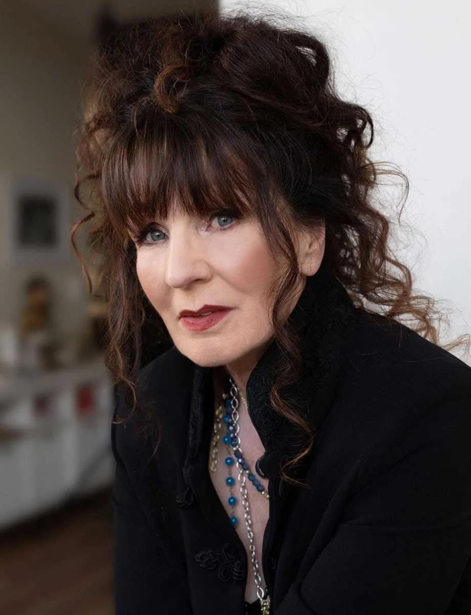 A woman with curly brown hair, blue eyes, wearing a black top and layered necklaces, posing indoors.