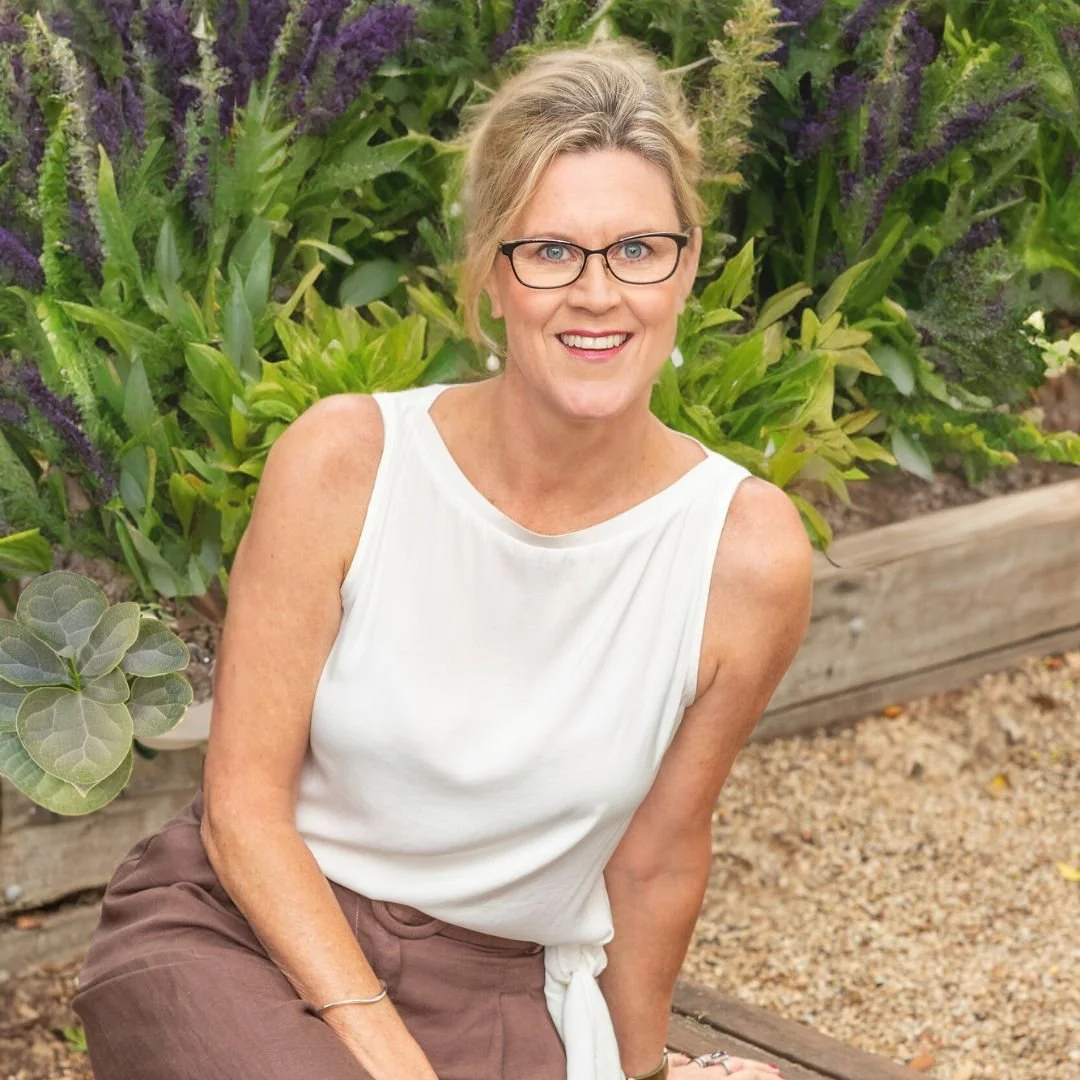 A smiling middle-aged woman with glasses, wearing a sleeveless white top and brown pants, sitting outdoors in front of green foliage and purple flowering plants.